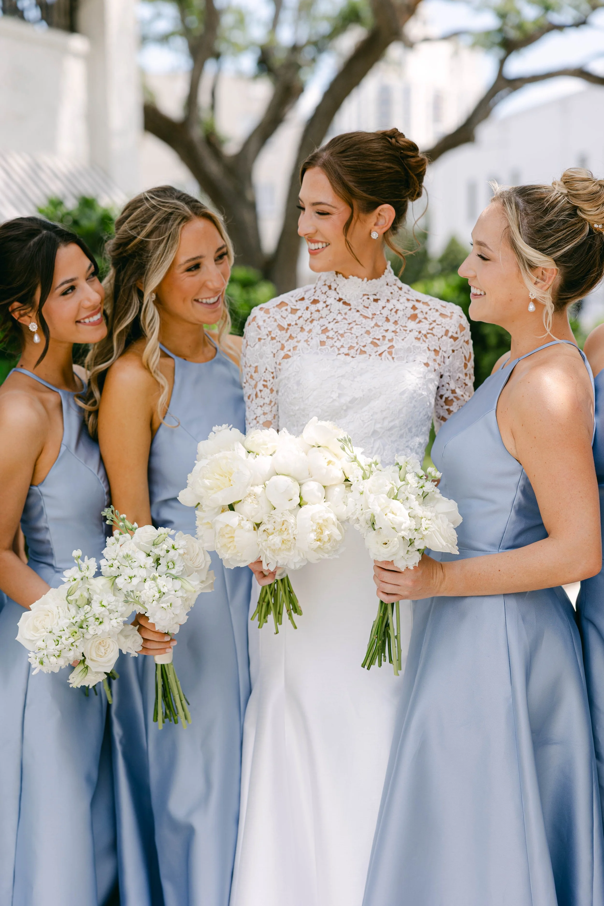 Women in wedding attire, including bride in white lace dress and bridesmaids in light blue dresses, holding white floral bouquets, outdoors under a tree.