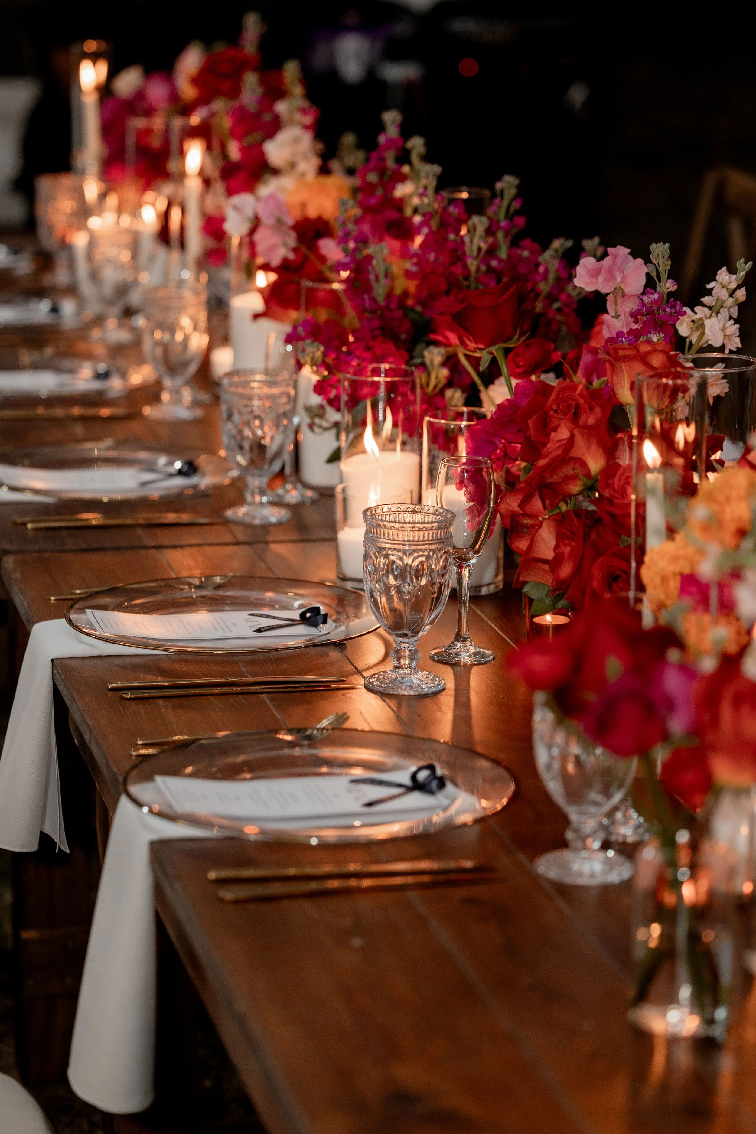 Elegant dinner table setup with floral centerpieces, candles, glassware, and plates with menus, in a dimly lit setting.
