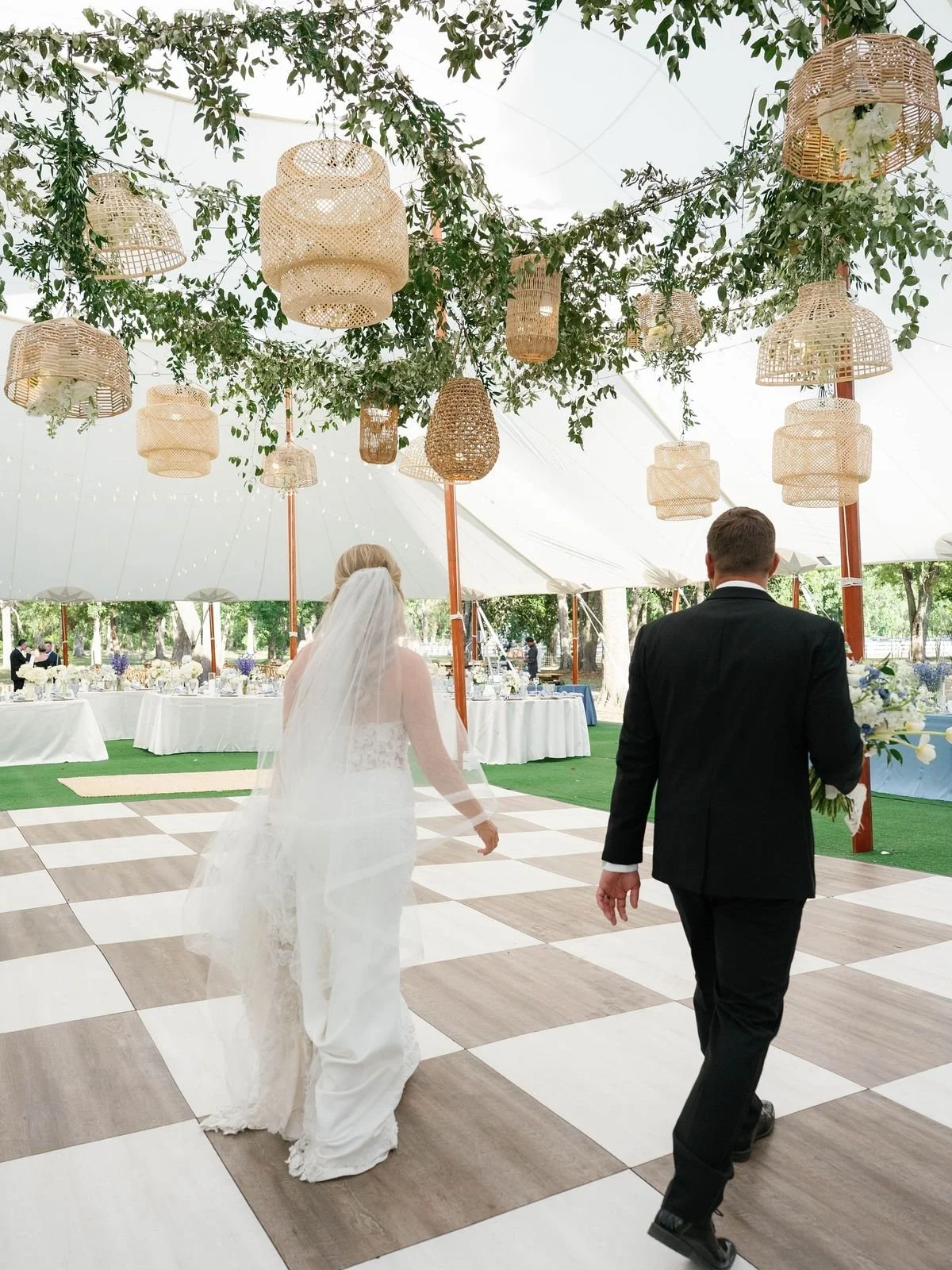 Bride and groom walking on a checkered dance floor at an outdoor wedding reception under hanging wicker lanterns and string lights, with decorated tables in the background.