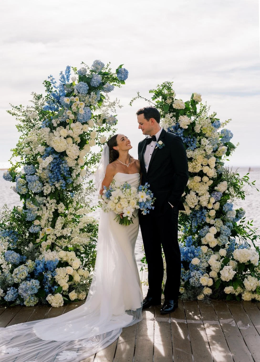 A bride and groom standing together on a beach, framed by a large floral arch of white and blue flowers, during their wedding ceremony.
