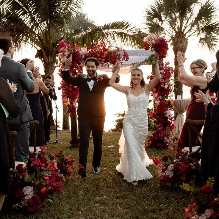 A newlywed couple, the groom in a tuxedo and the bride in a white lace wedding dress, walking down the aisle at an outdoor wedding ceremony during sunset, surrounded by family and friends, with palm trees and floral decorations.