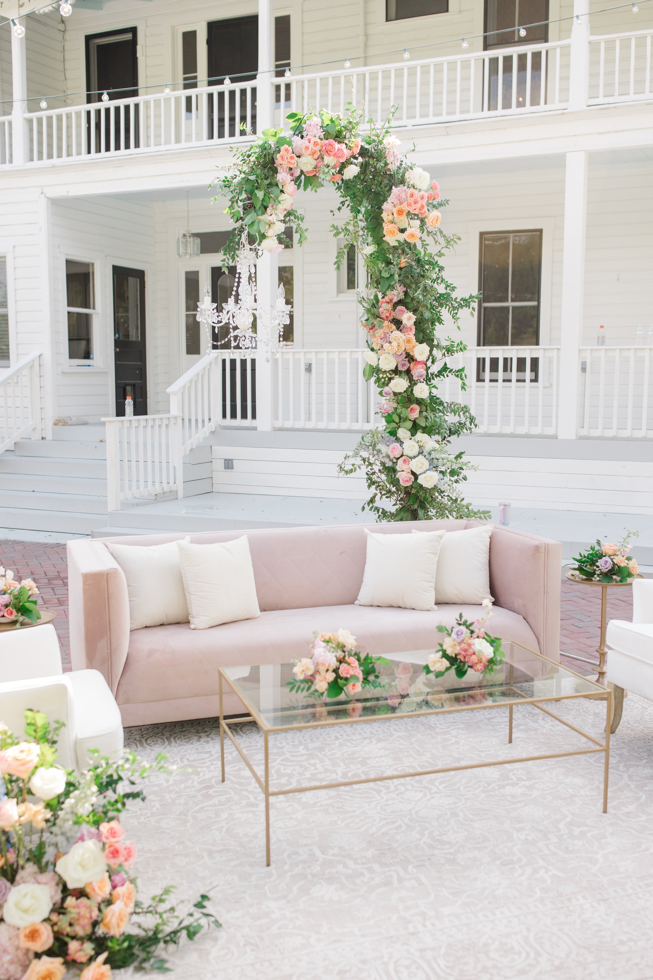 Indoor wedding or event setup with a blush pink sofa, glass coffee table with flower arrangements, and large floral arch with pink and white roses and greenery.