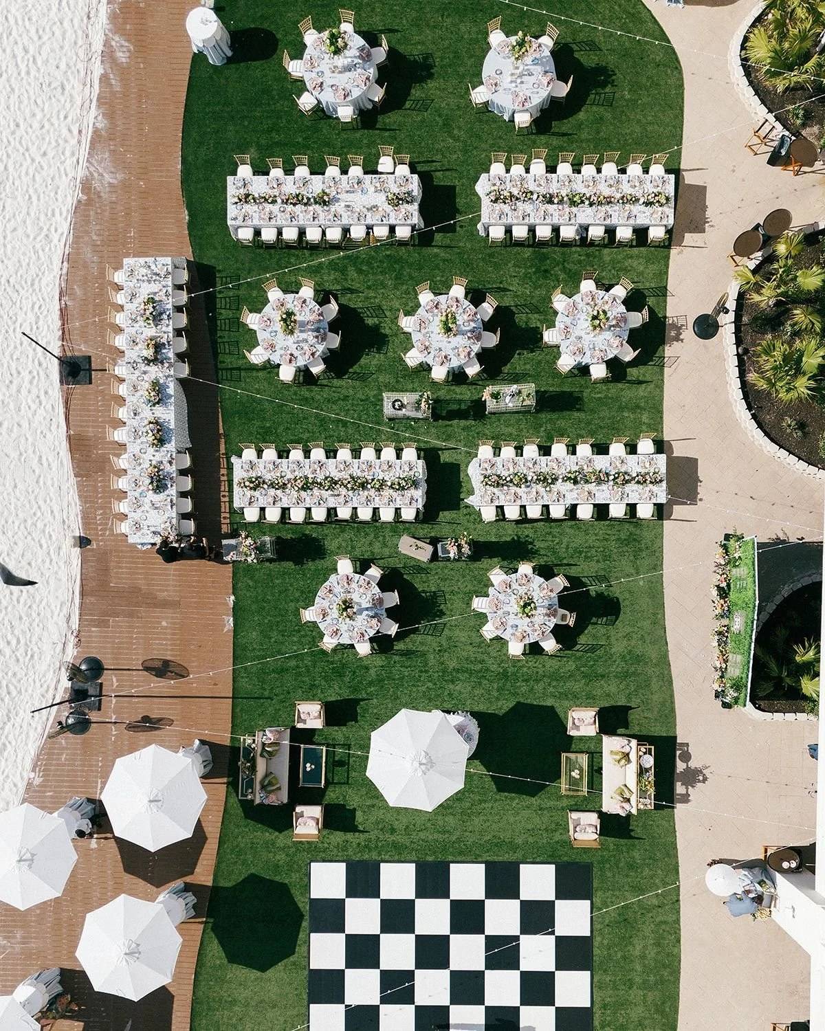 Aerial view of an outdoor event setup with round and long tables decorated with floral centerpieces, umbrellas, and a checkered dance floor surrounded by white lounge furniture on green grass.