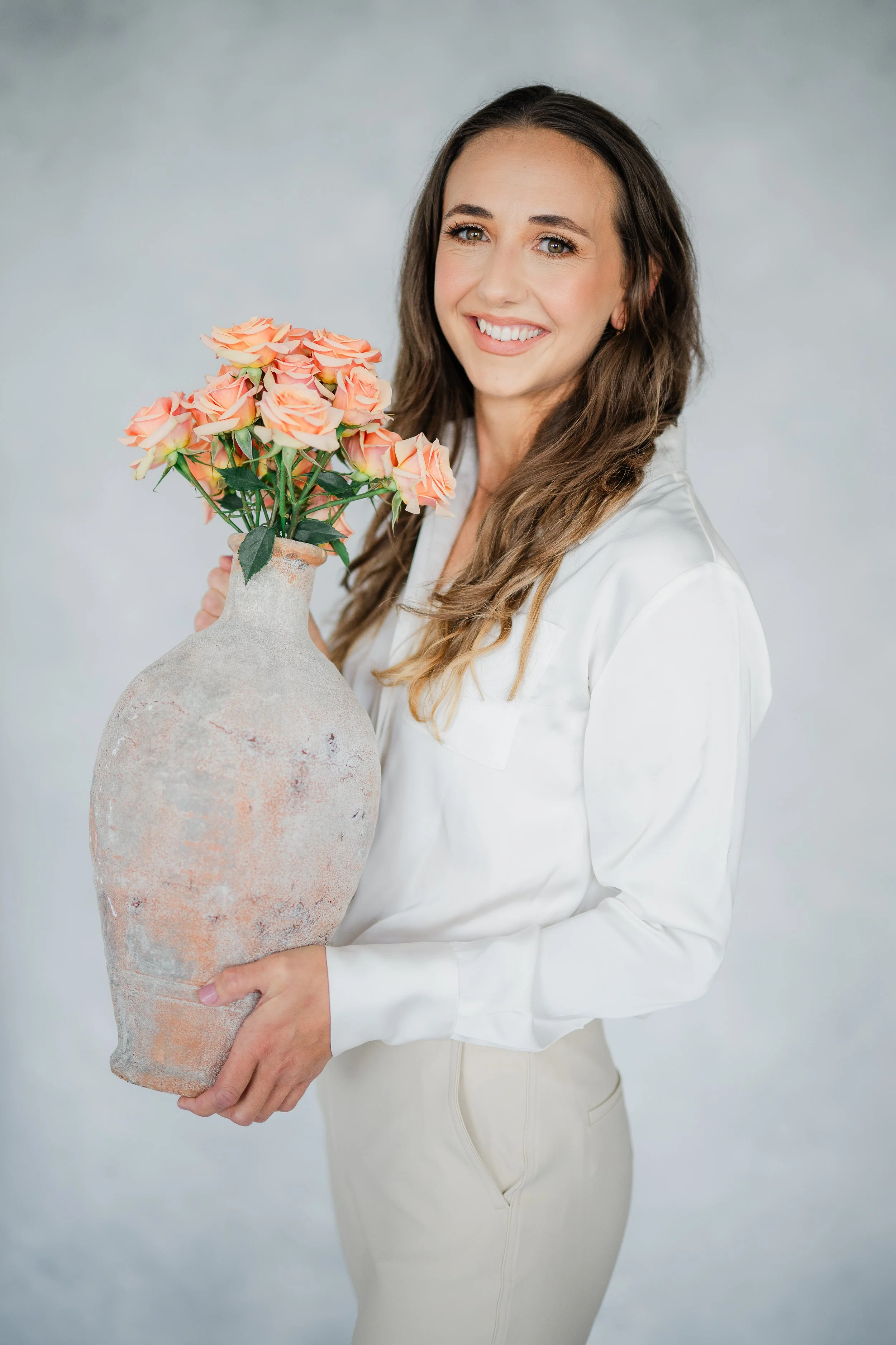 A woman with long brown hair holding a large, textured ceramic vase filled with peach-colored roses, smiling at the camera against a plain light gray background.