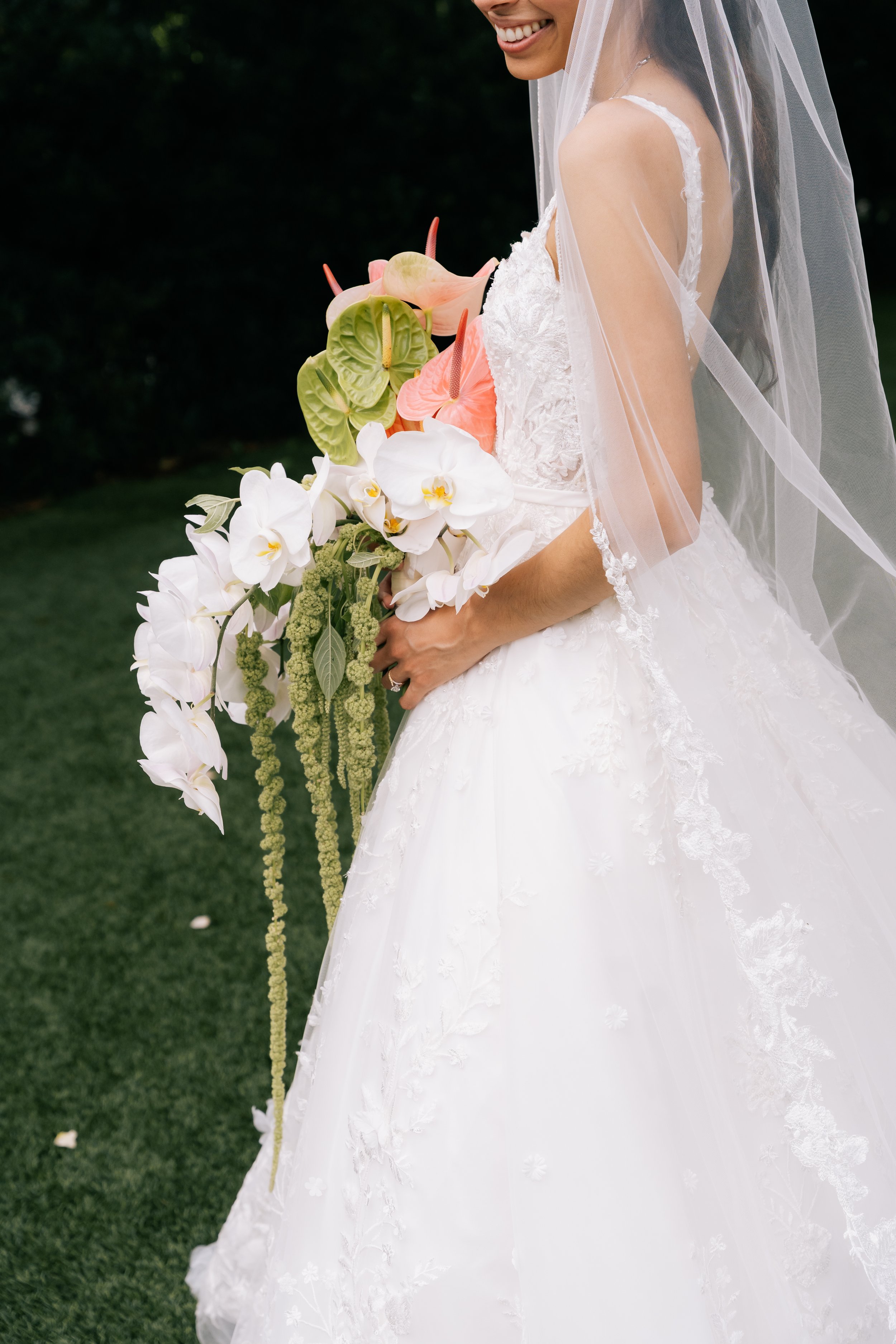 Close-up of a smiling bride holding a large bouquet of white and pink flowers with green and pink foliage, wearing a white lace wedding dress and veil outdoors.