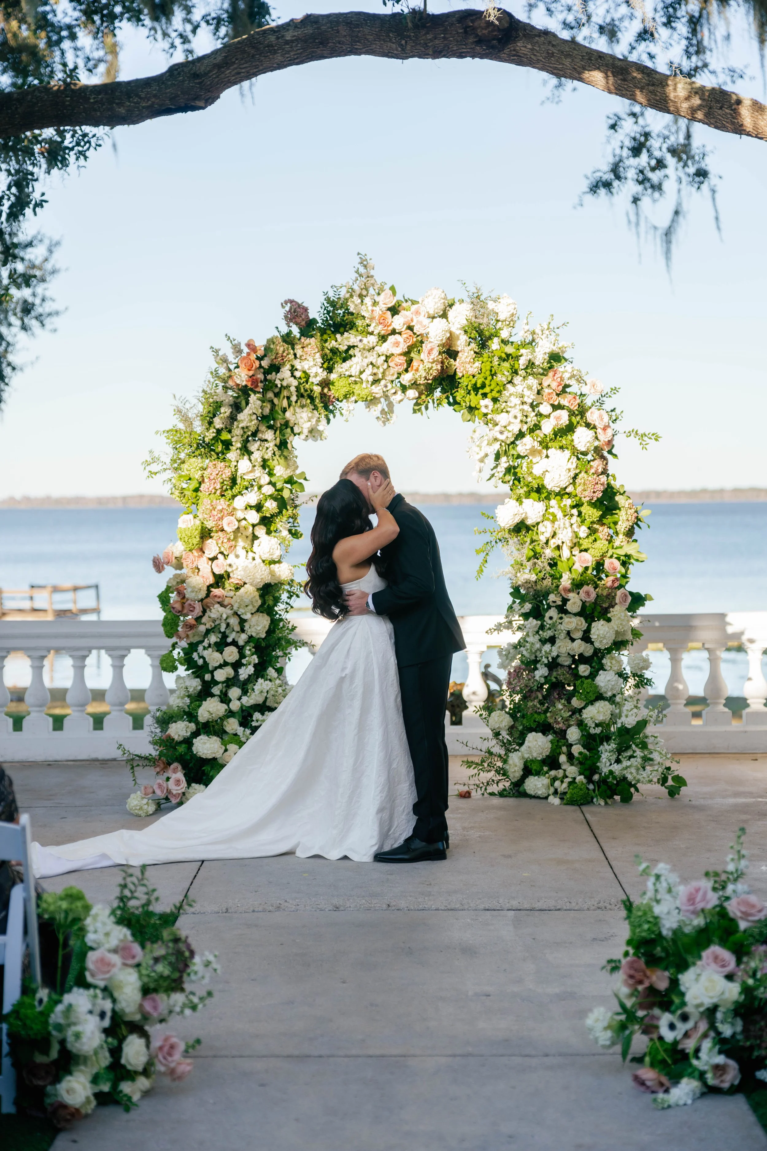 A bride and groom sharing a kiss under a floral arch on a lakeside outdoor wedding ceremony.