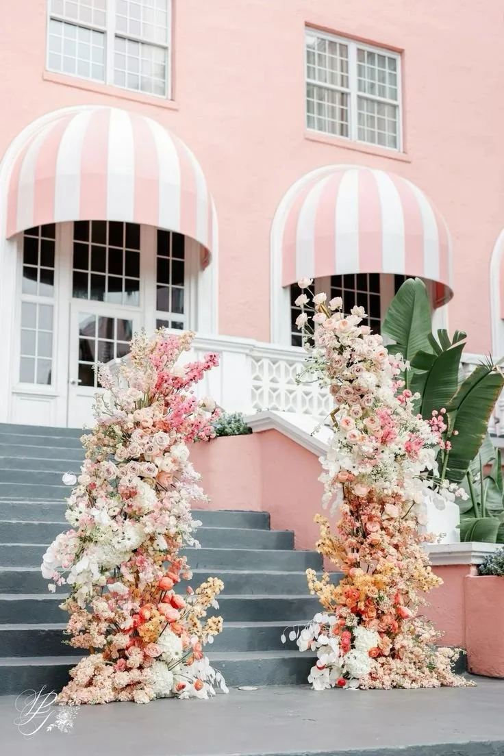 Flower arch with pink and white blooms on gray steps outside a pink building with striped pink and white awnings.