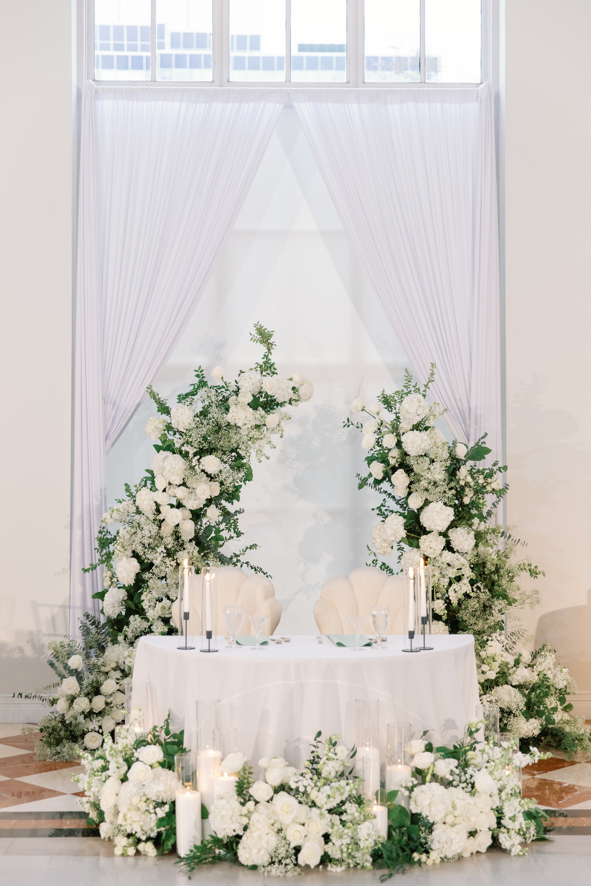 Elegant wedding reception setup with a white round table, floral arrangements of white flowers and greenery, and tall white candles in glass holders, positioned in front of a large window with sheer white curtains and a display of white flowers at the base.