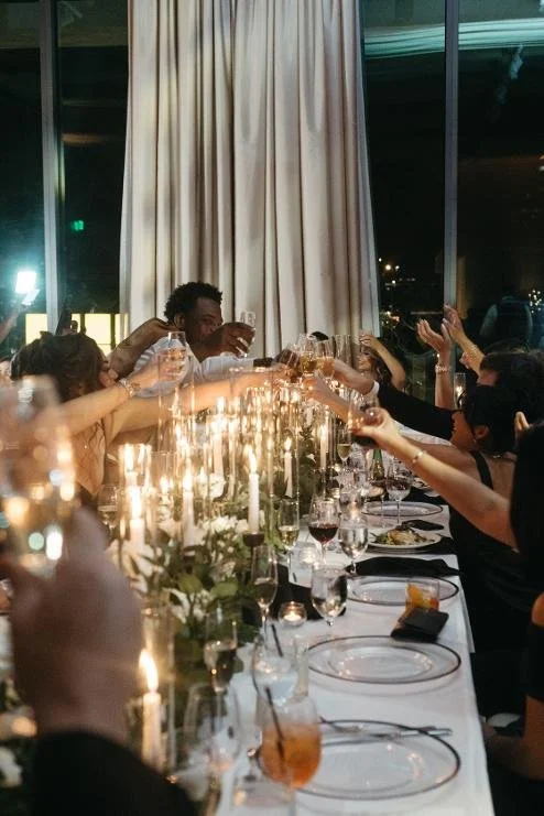 Group of people celebrating at a formal dinner party, raising glasses in a toast, with candlelit table, wine glasses, dinner plates, and floral centerpieces.
