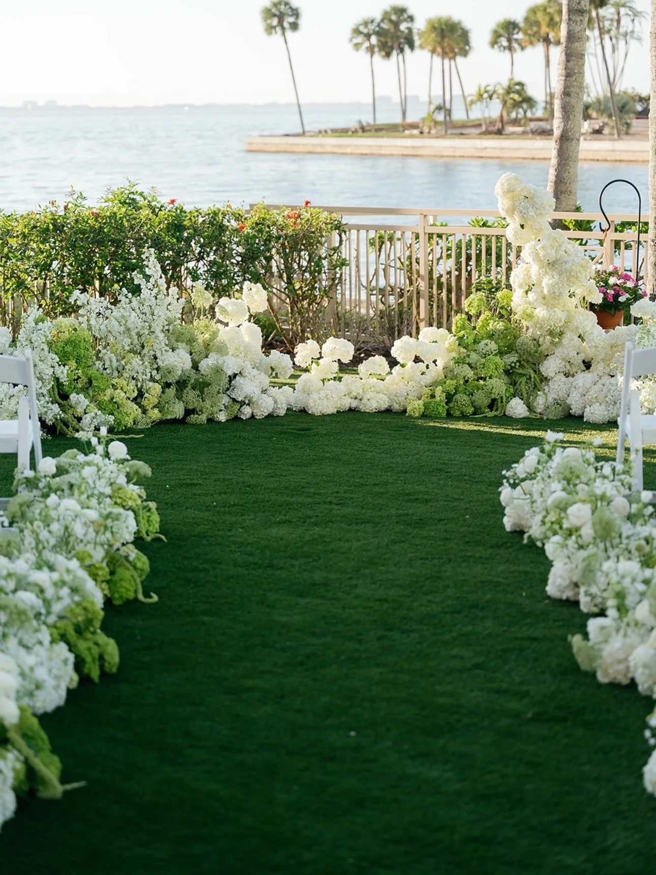 A decorated outdoor wedding aisle along a waterfront, with white and green floral arrangements lining a green carpet, and a backdrop of water, palm trees, and a distant shoreline.