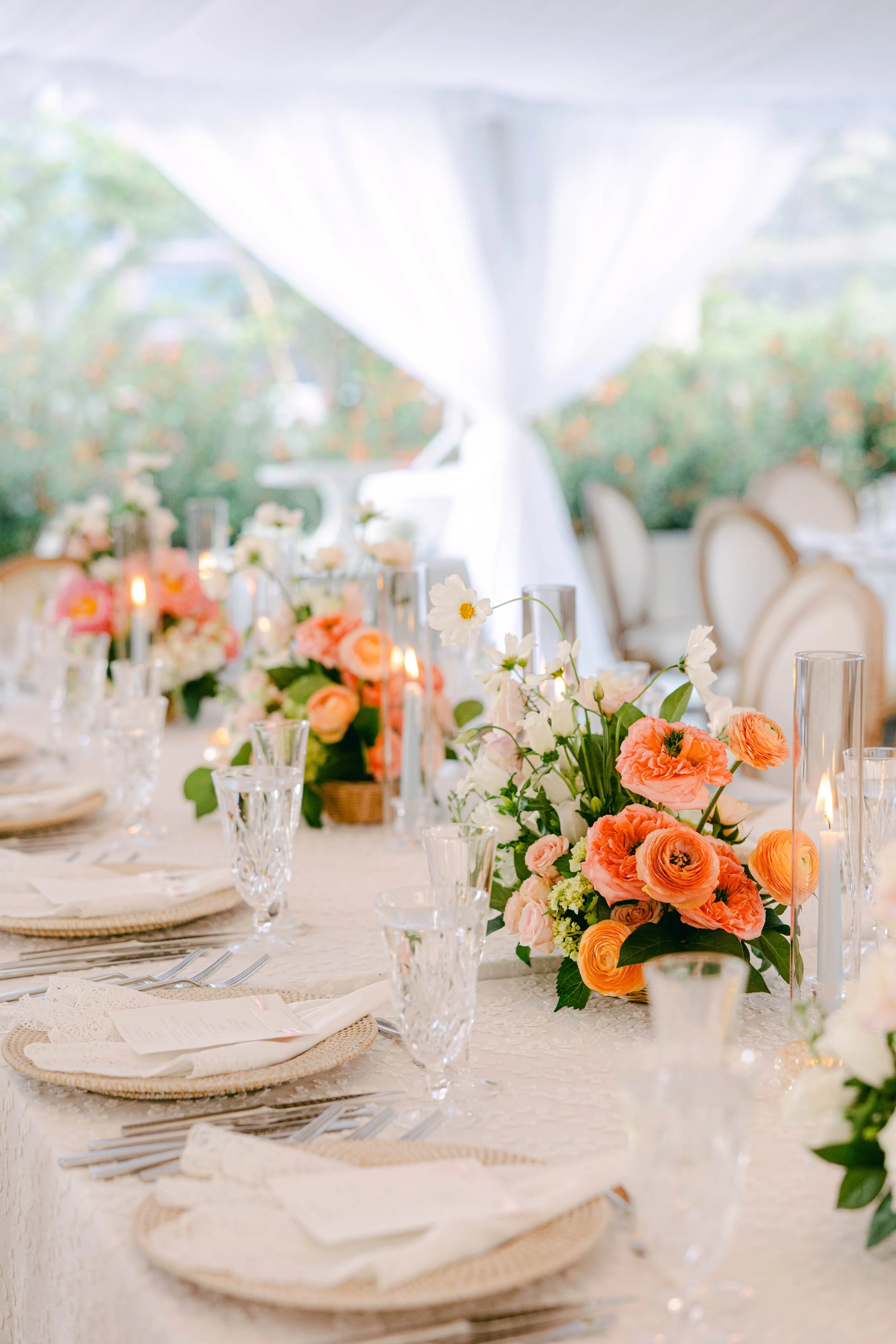 A decorated dining table with floral centerpieces, candles, glassware, and place settings under a white tent at an outdoor event.