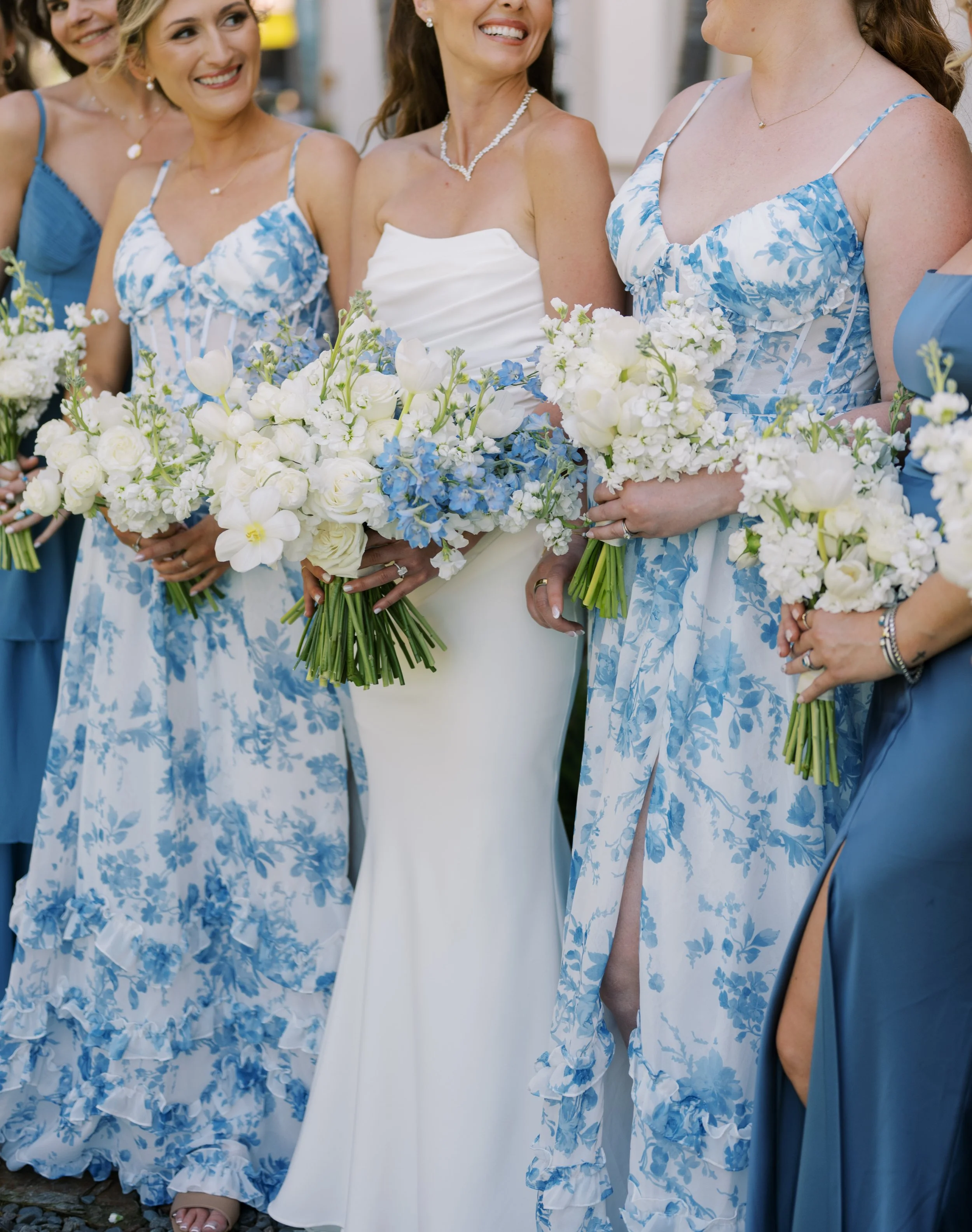 A group of women dressed in blue and white floral dresses standing close together outdoors, holding bouquets of white and blue flowers, smiling, at what appears to be a wedding or celebration.