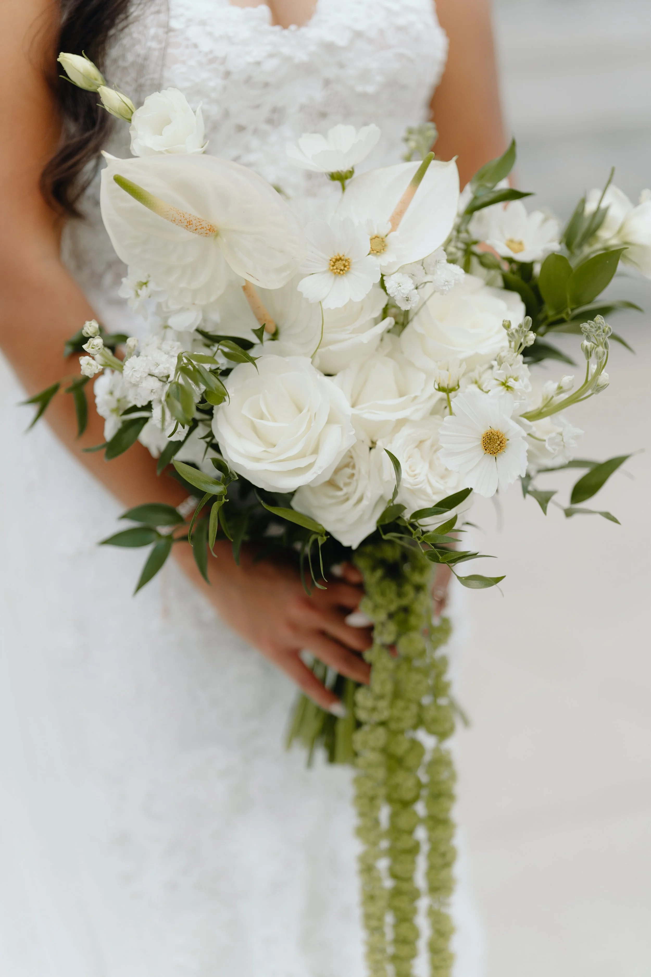 Close-up of a bride holding a bouquet of white flowers, including roses, lilies, and small white blossoms, with a blurred background.