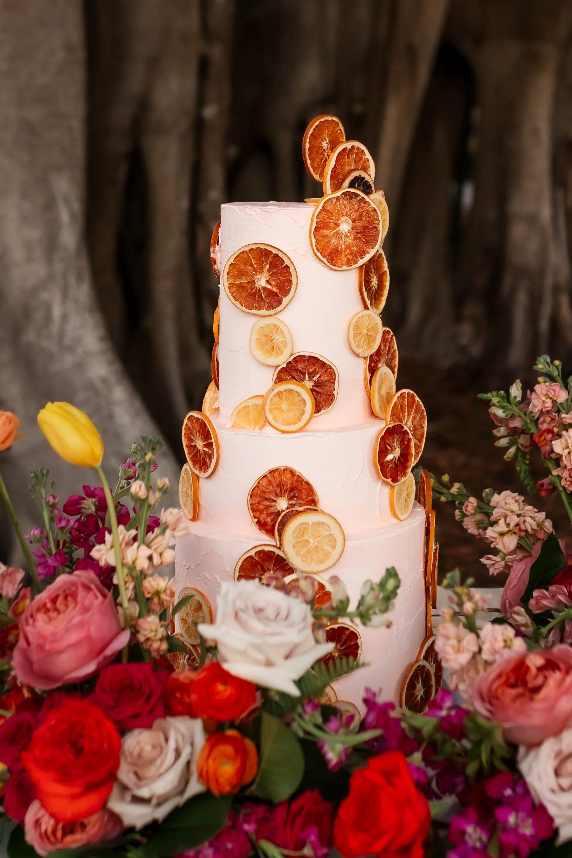 A tall white wedding cake decorated with slices of dried citrus fruits, surrounded by colorful flowers including roses and other blossoms.
