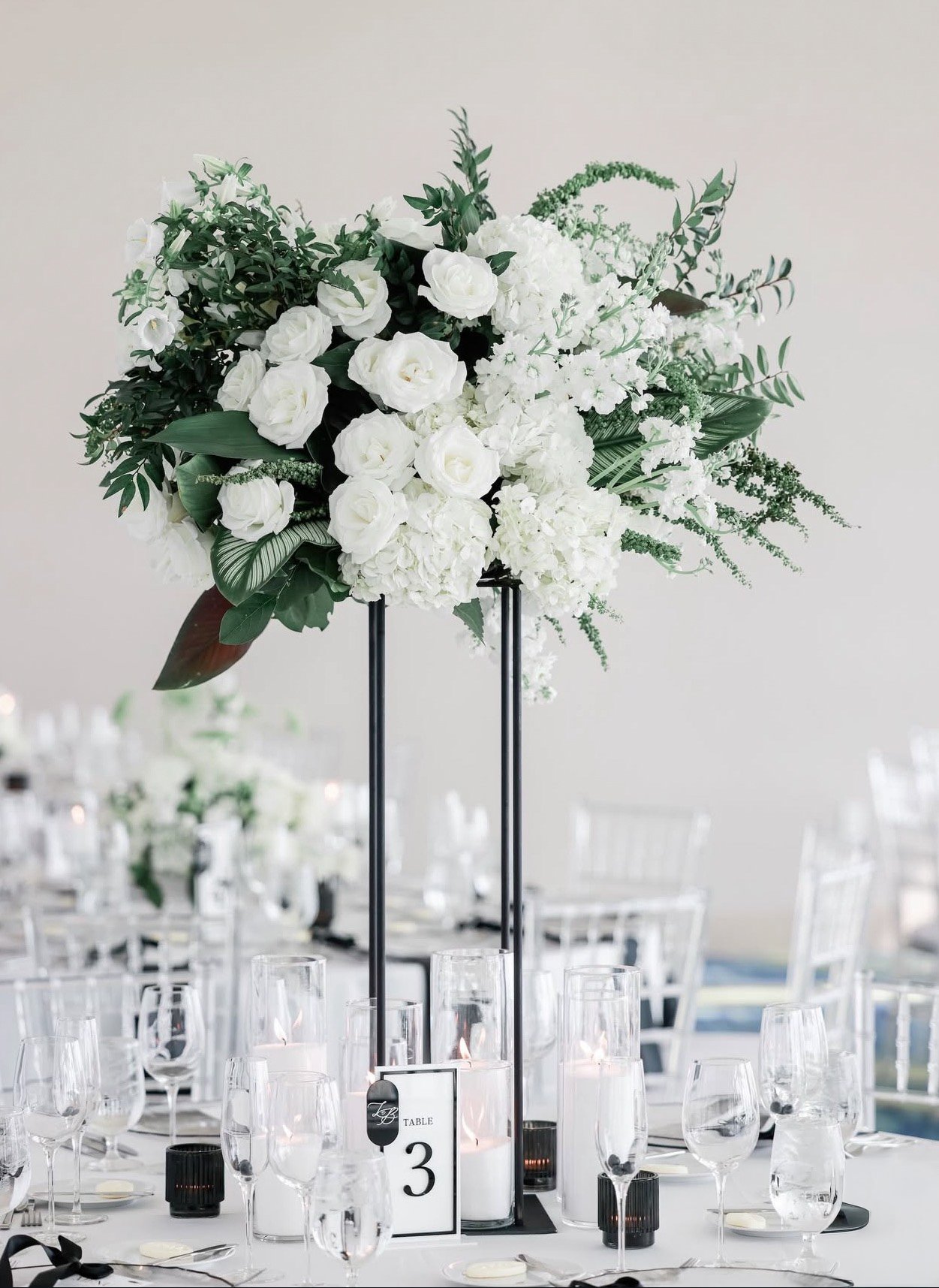 A tall floral centerpiece with white roses, hydrangeas, and greenery on a table at a formal event, with glassware and candles around it.