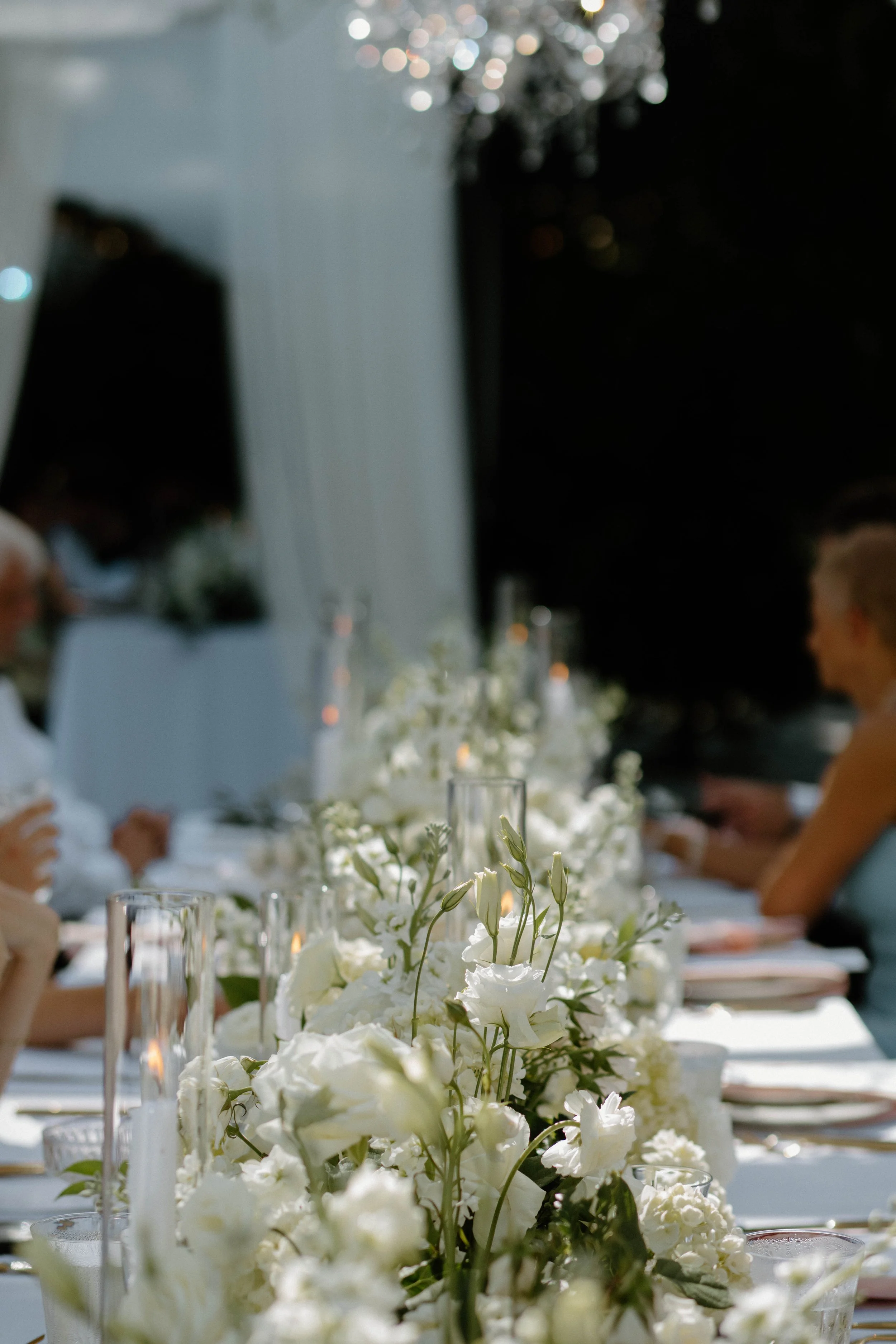 Close-up of a floral centerpiece featuring white flowers on a banquet table at a formal event, with a blurred background of guests, candles, and fairy lights.
