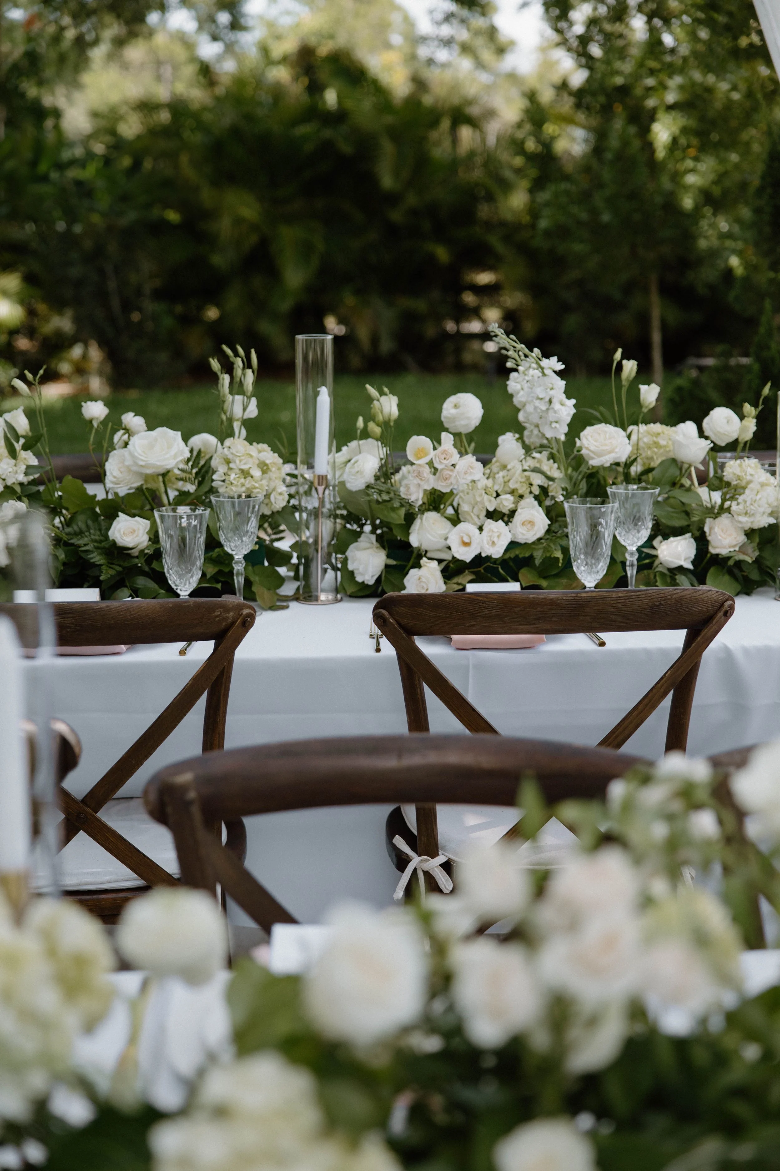 A beautifully decorated outdoor table with white flowers, glassware, and a tall candle holder, set amidst a lush green garden.