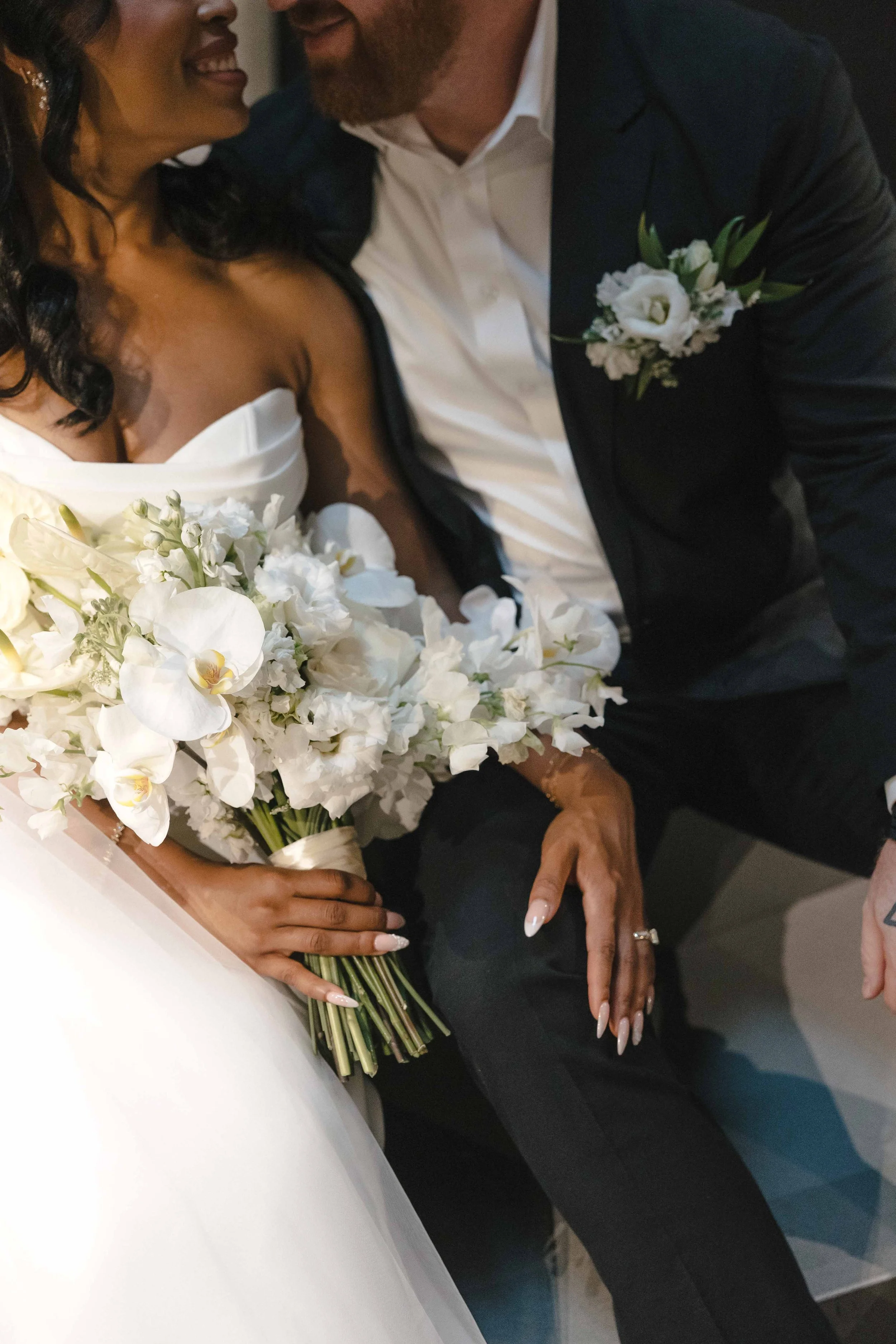 Close-up of a couple at a wedding, showing the woman in a white strapless wedding dress holding a bouquet of white flowers and the man in a black suit with a white shirt, with their faces close together.
