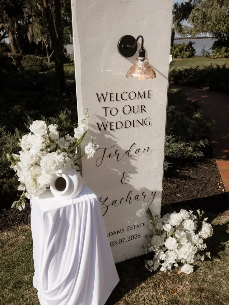 Wedding welcome sign with floral decorations and a white draped table outside.