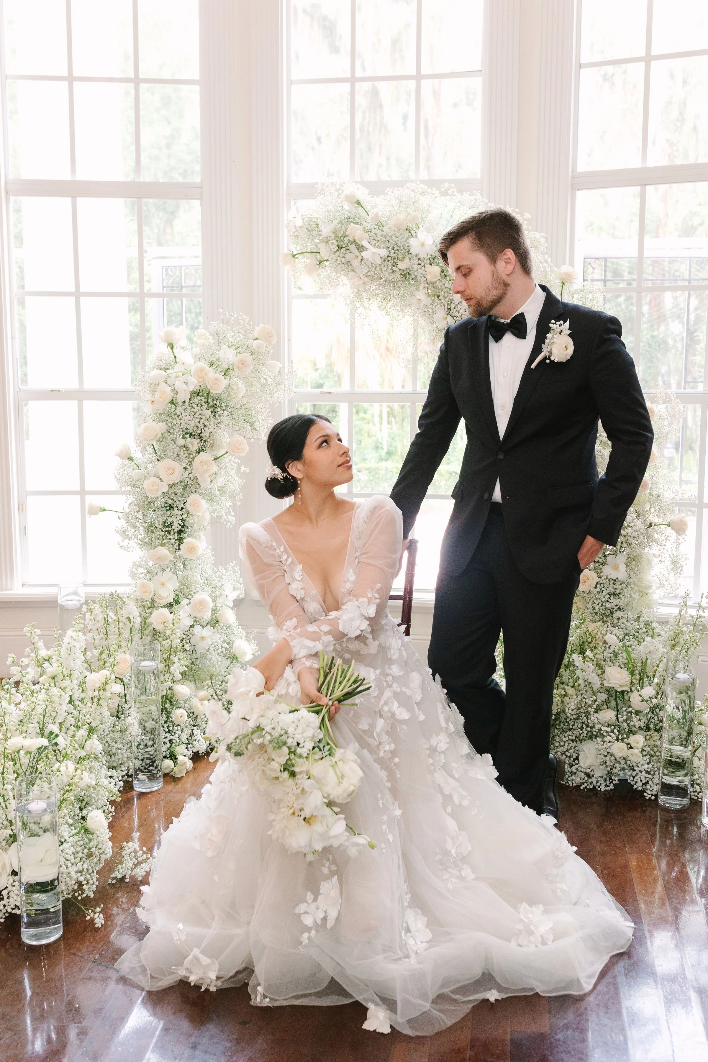 A bride in a white wedding gown sitting and holding a bouquet of white flowers, looking up at a groom in a black tuxedo with a bow tie, in a room decorated with white flowers and large windows.