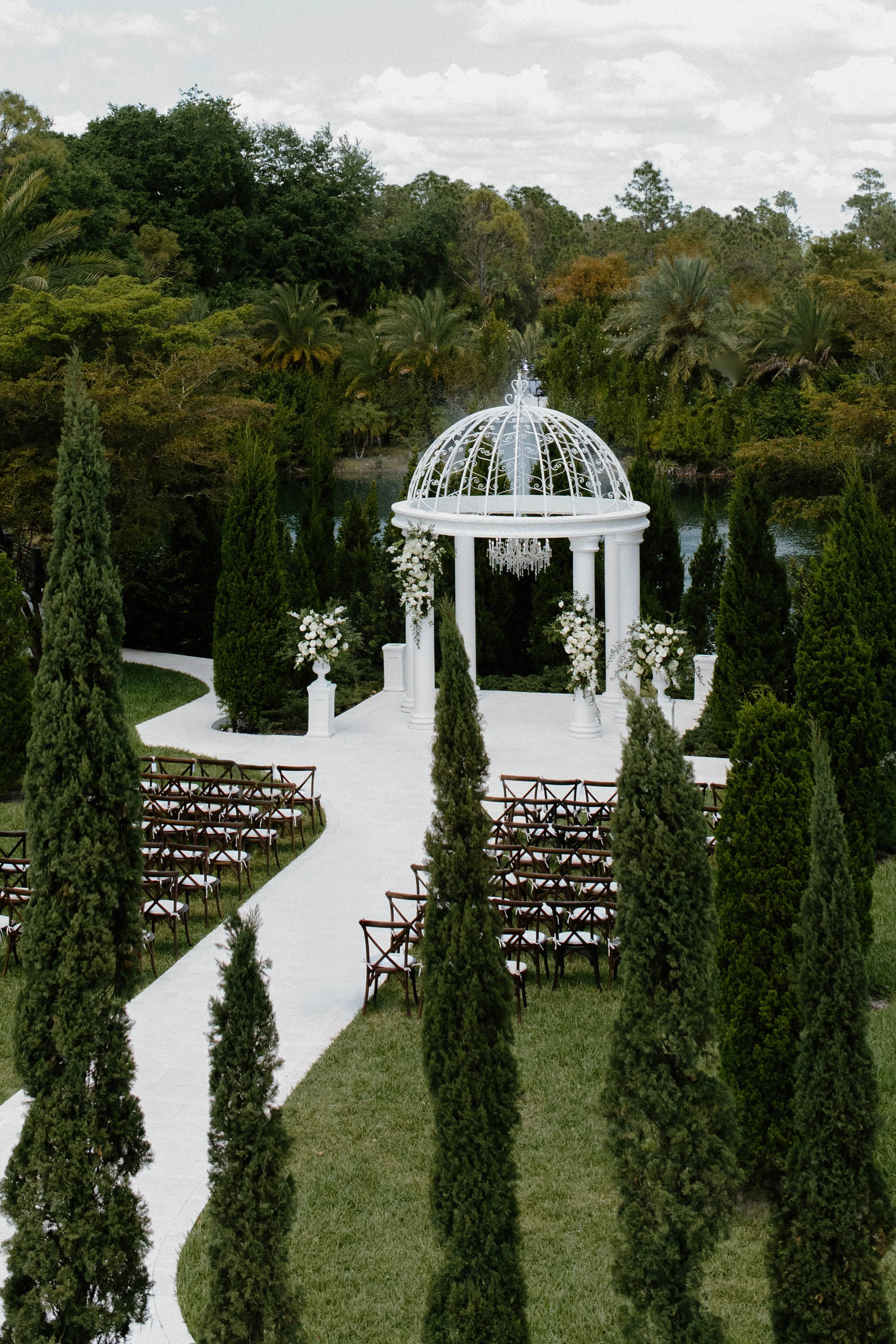A white, wrought-iron gazebo with floral decorations stands at the end of a white aisle, surrounded by tall green trees and chairs for an outdoor wedding ceremony.