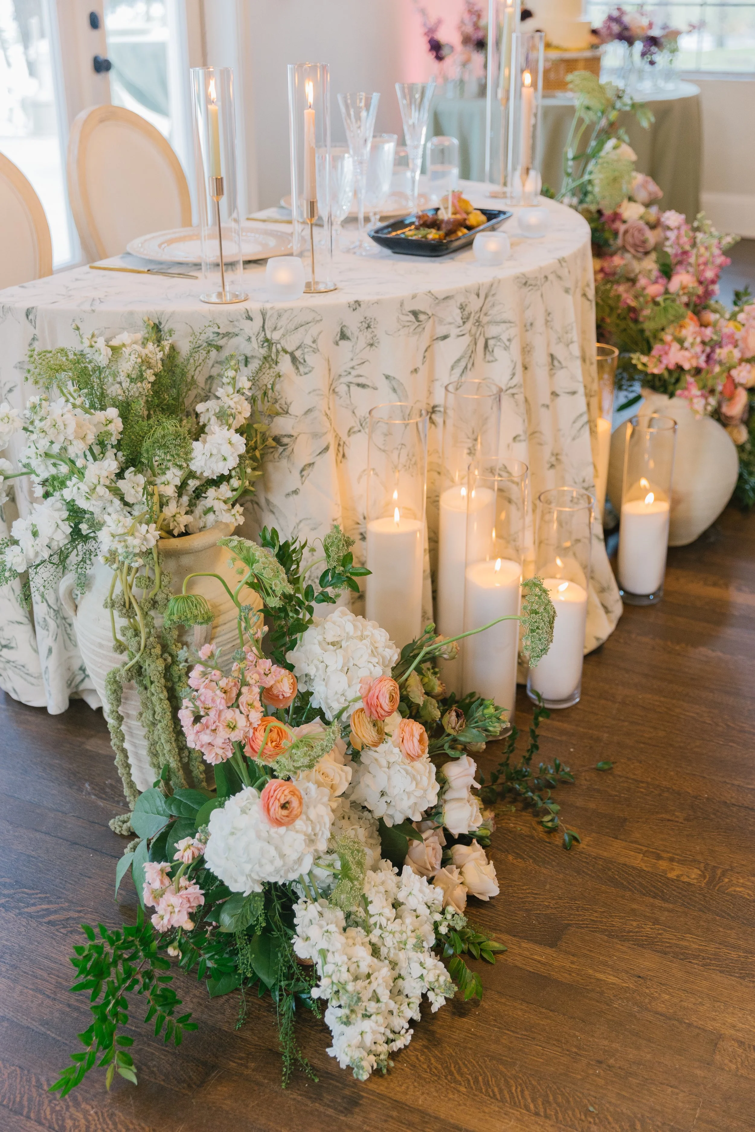 A decorated table with candle holders, floral arrangements, and a floral tablecloth, surrounded by flowers and candles on a wooden floor, likely set for an event or celebration.