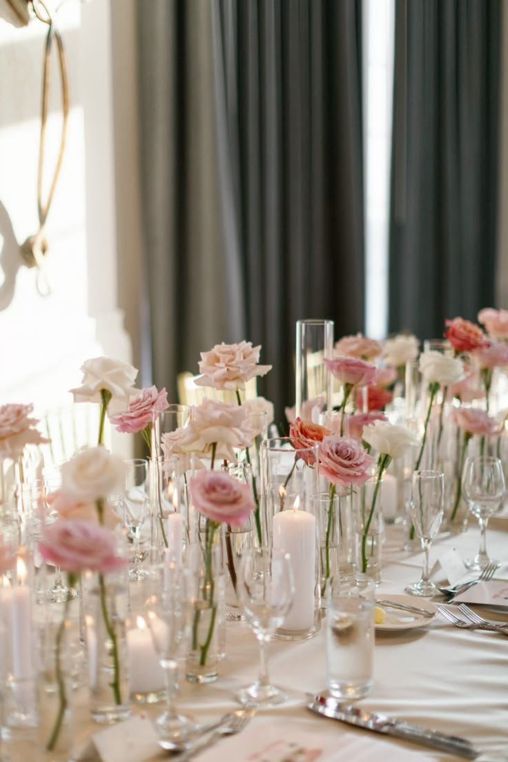A Wedding reception table decorated with pink and white flowers in tall glass vases, white candles, and place settings for a formal event, with dark curtains in the background.
