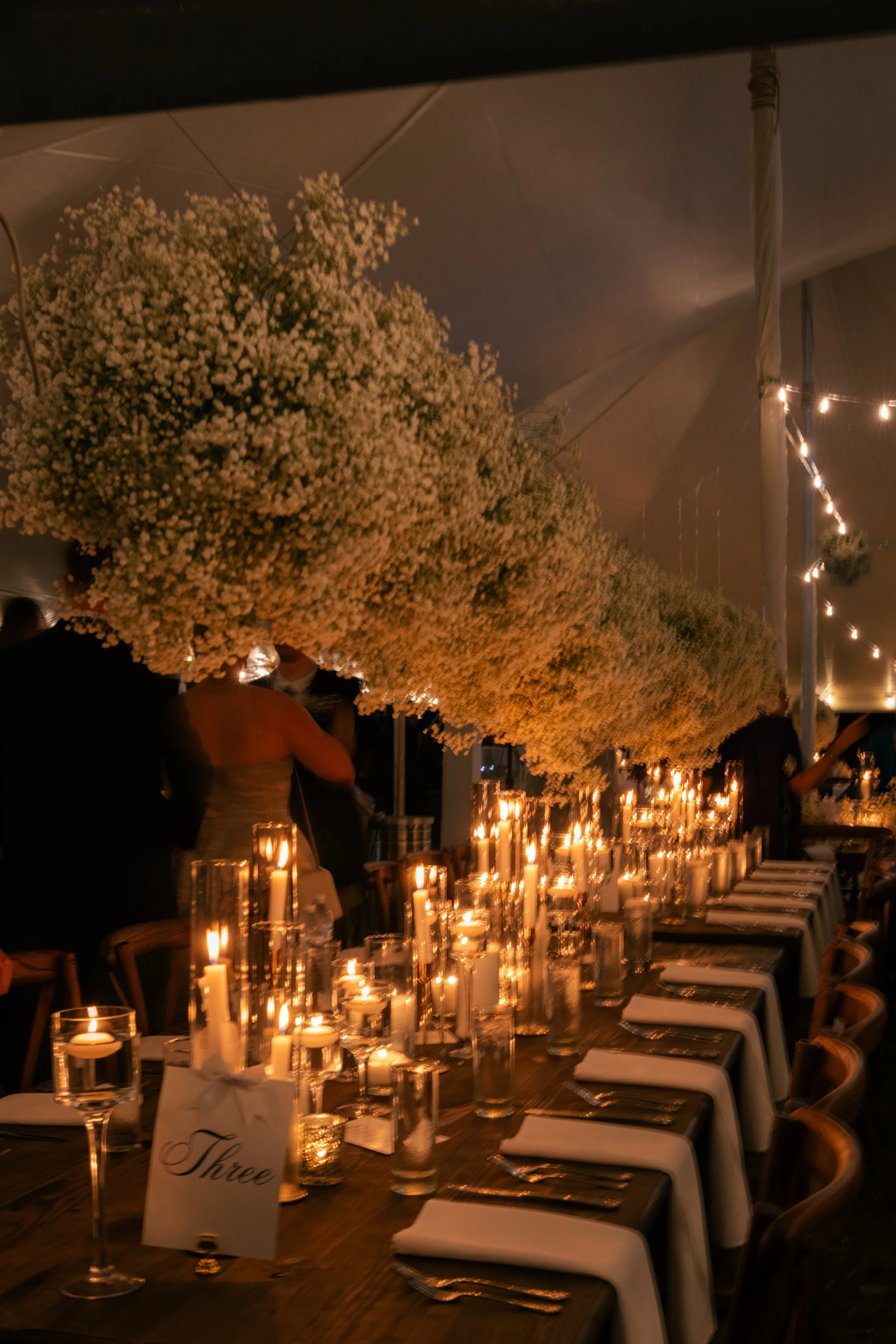 Long dinner table decorated with numerous lit candles in glass holders, surrounded by chairs. Overhead, large arrangements of white baby's breath flowers decorate the ceiling, with warm string lights hanging beside them. A small card on the table reads 'Three'.