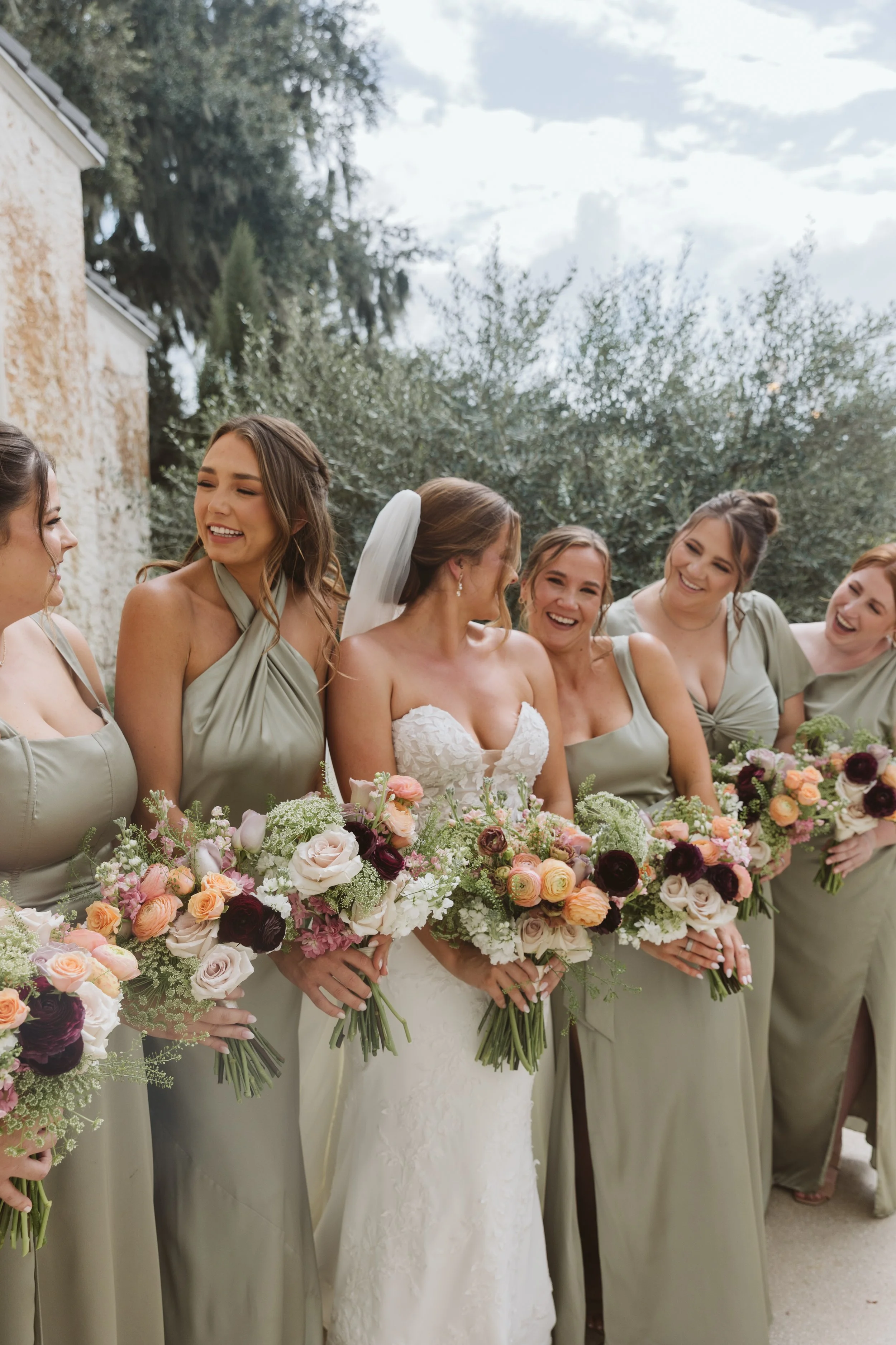 A bride and her bridesmaids standing together outdoors, holding bouquets of flowers, during a wedding celebration in daytime.