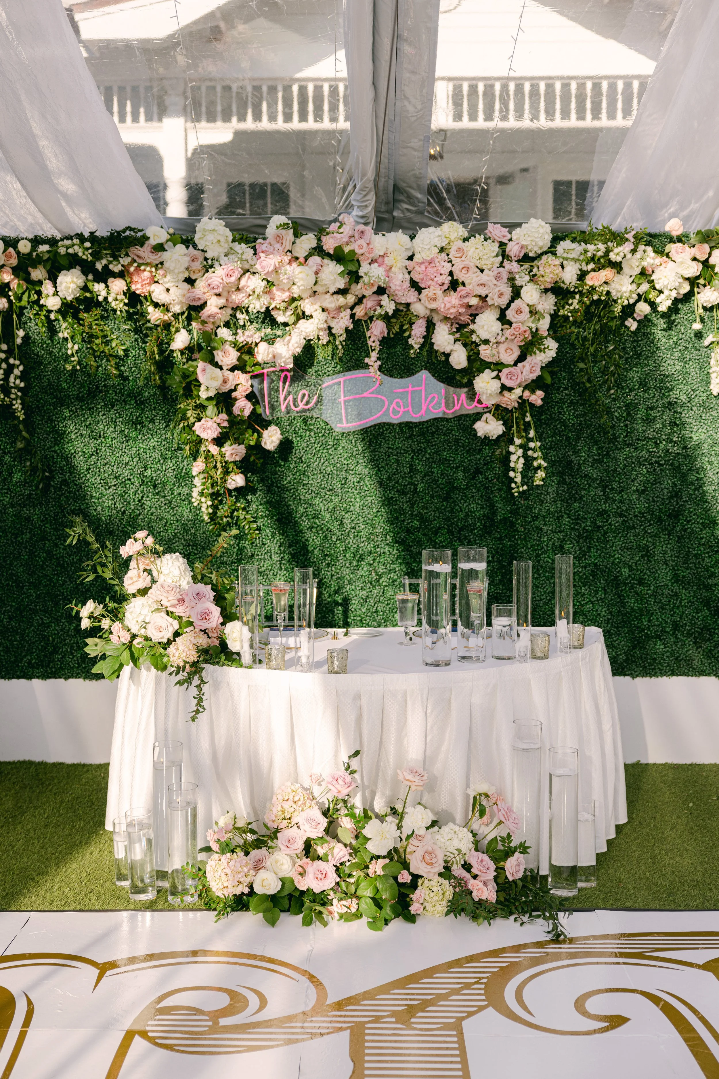 A decorated sweetheart table at a wedding reception with a backdrop of lush green foliage and a window with curtains. The table has a white tablecloth, surrounded by glass candles and a large floral arrangement of white and pink flowers.