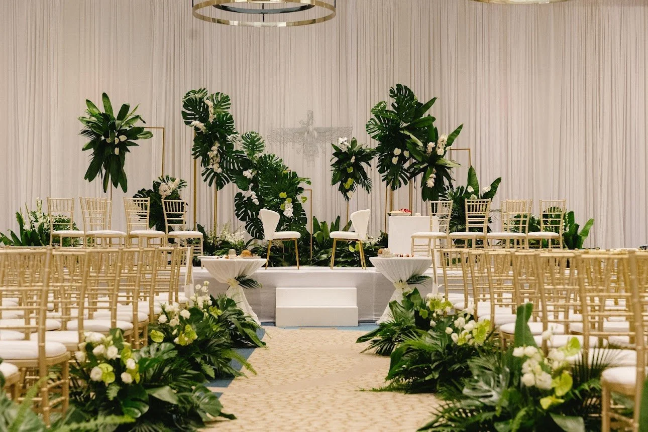 Wedding ceremony setup with gold chairs, white cushioned seats, floral arrangements, and a small stage with two white chairs and greenery background.