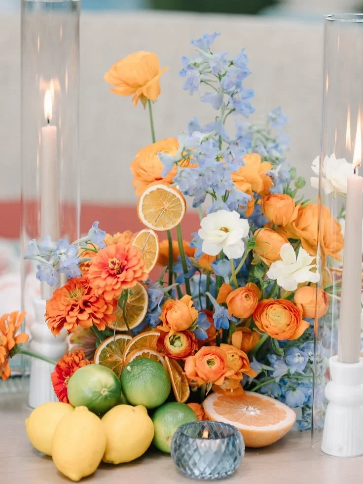 Colorful floral arrangement with orange, white, and blue flowers, lemons, limes, and grapefruit, flanked by tall white candles in glass holders.