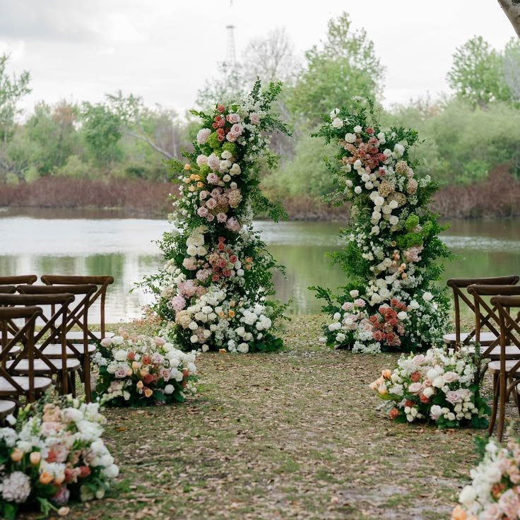 Outdoor wedding ceremony setup with a floral arch by a lake, adorned with pink and white flowers and greenery, flanked by wooden chairs.