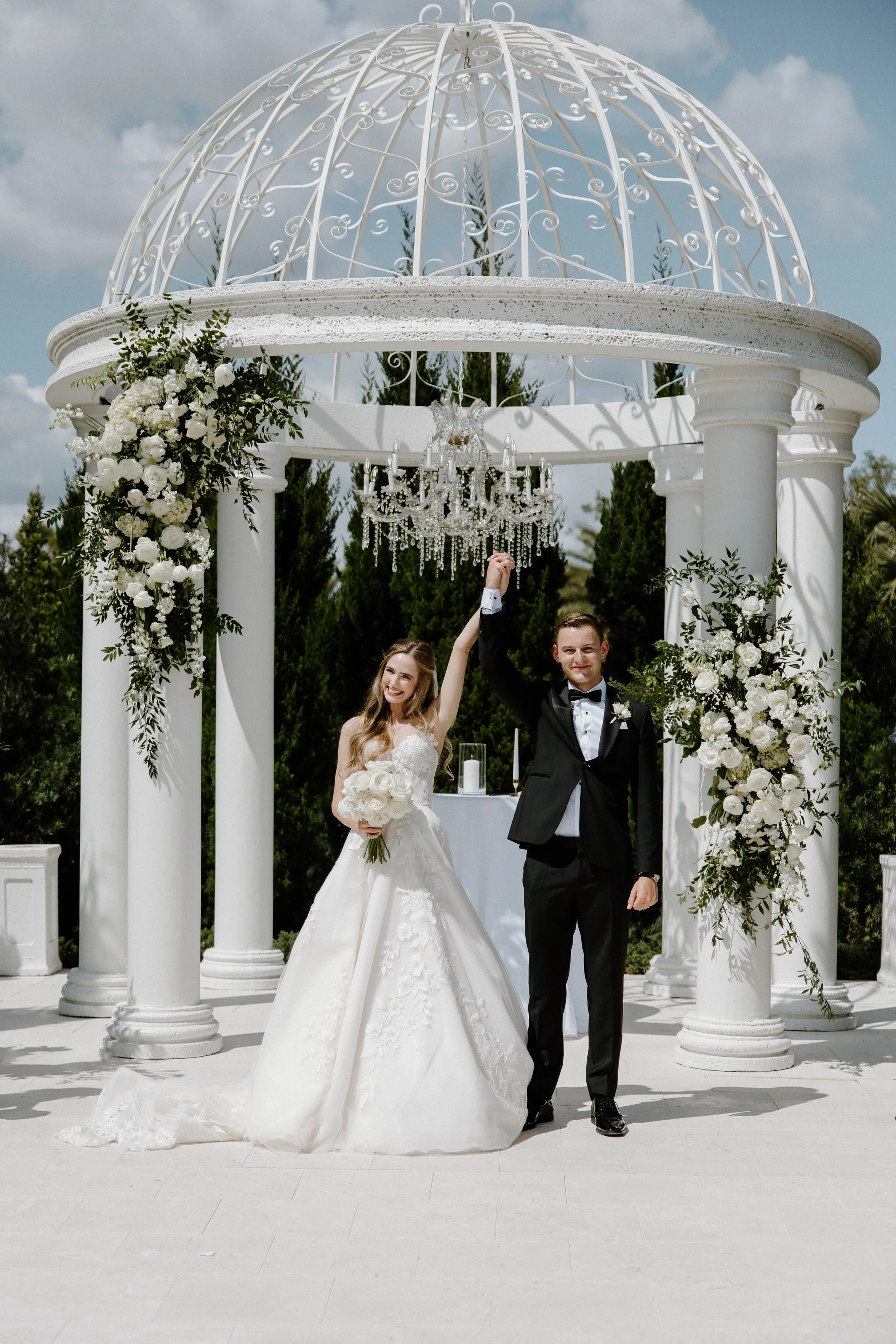 A newlywed couple standing under a white ornate gazebo decorated with white flowers and greenery, with a chandelier hanging above them. The bride is holding a bouquet and wearing a white wedding gown, and the groom is wearing a black tuxedo. They are holding hands and smiling.