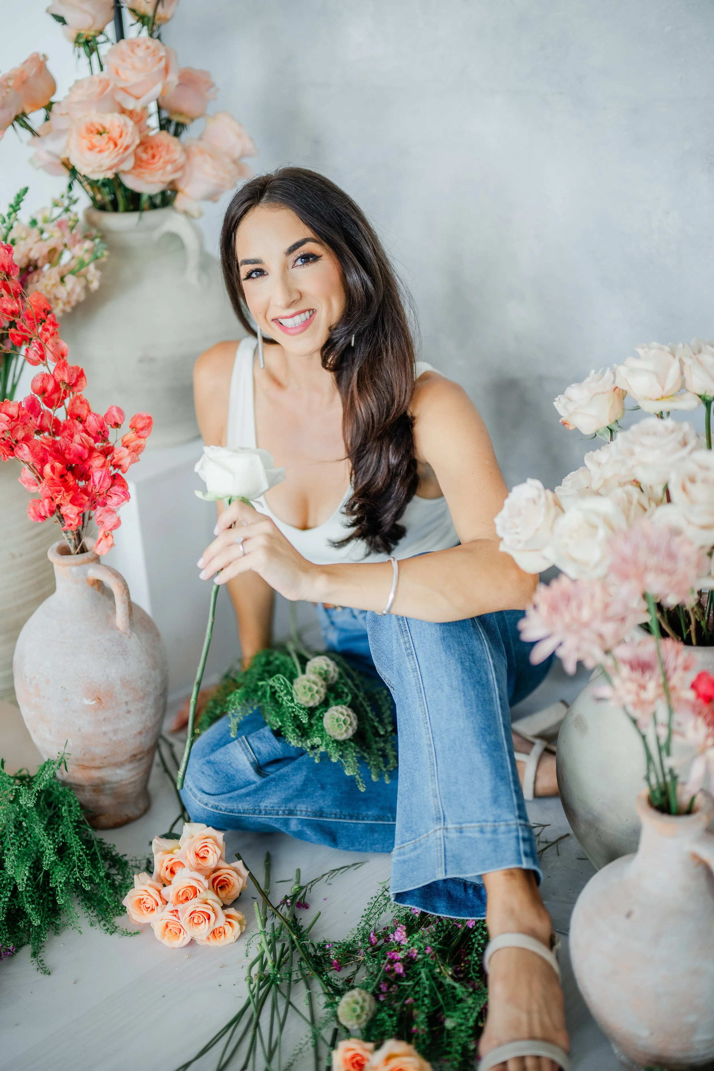 A woman with long dark hair smiles while sitting among various pink, peach, and red flowers in vases and scattered on the floor, with a neutral gray wall in the background.