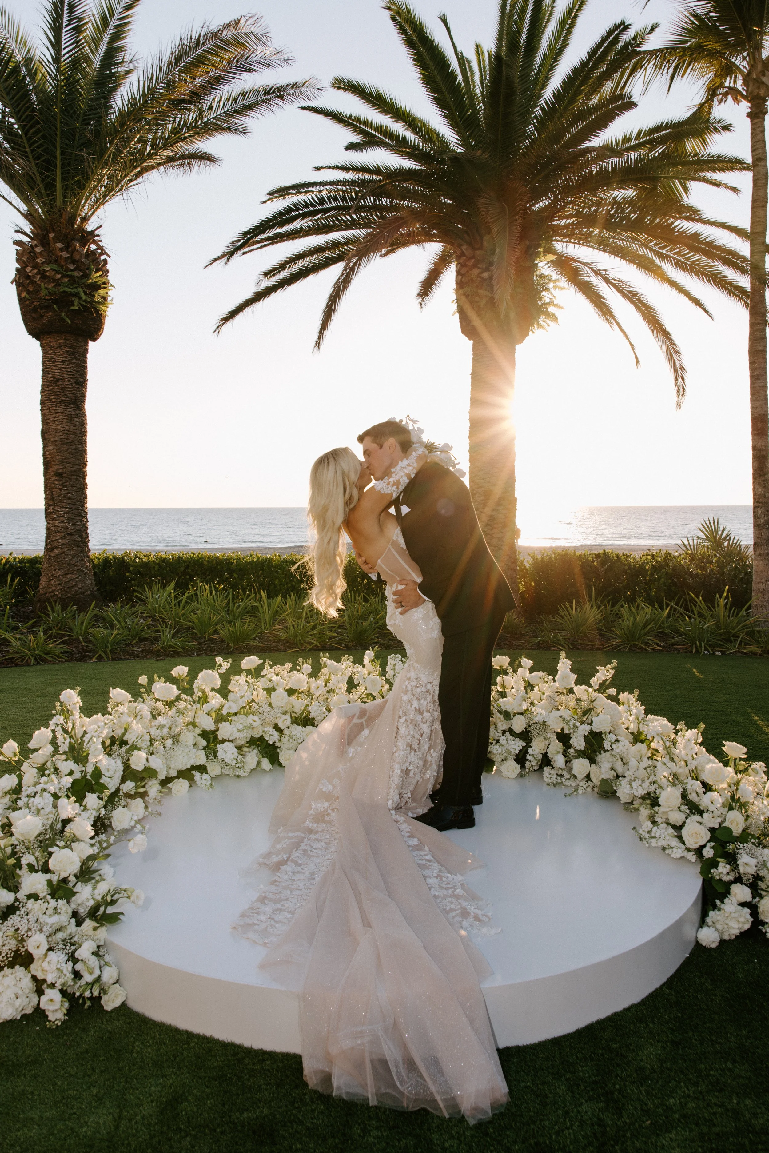 A bride and groom kissing during their wedding ceremony outdoors near the ocean, with palm trees in the background, sunlight shining through, and a floral arch decorated with white flowers.