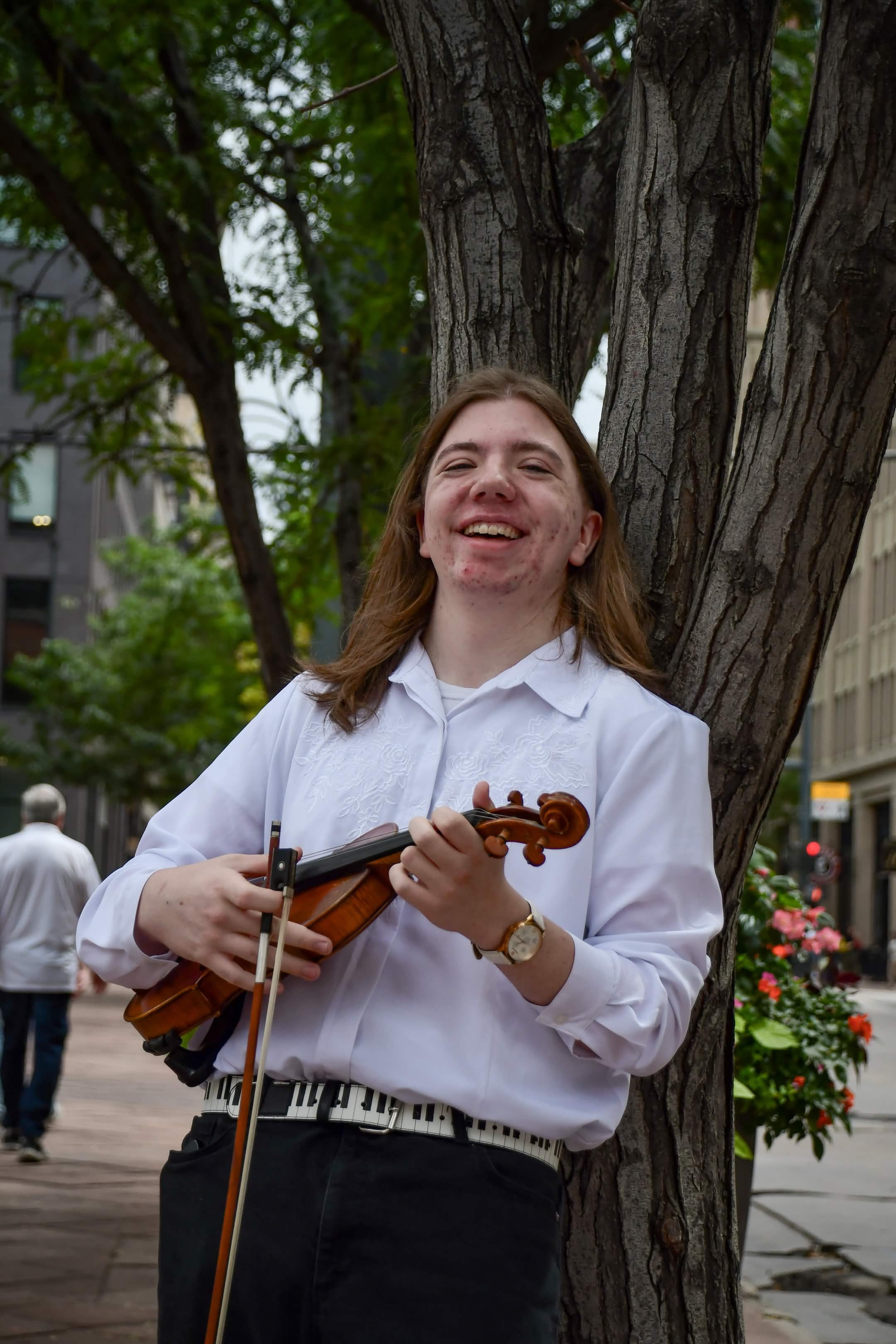 A person playing the violin outdoors in an urban setting, standing beside a tree, with buildings and people in the background.