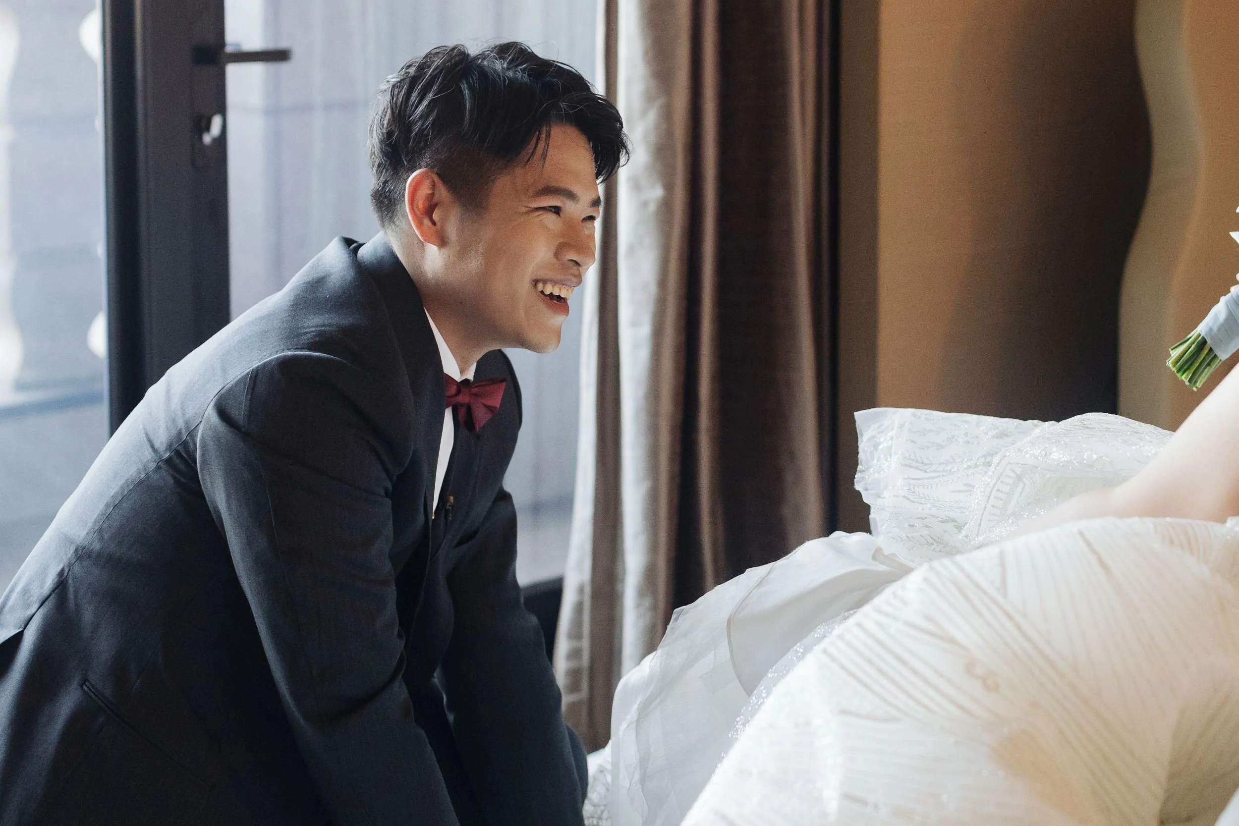 A man in a black suit with a red bow tie is smiling and leaning over a bed, looking at a person lying on it, with a background of a room with curtains and a glass door.