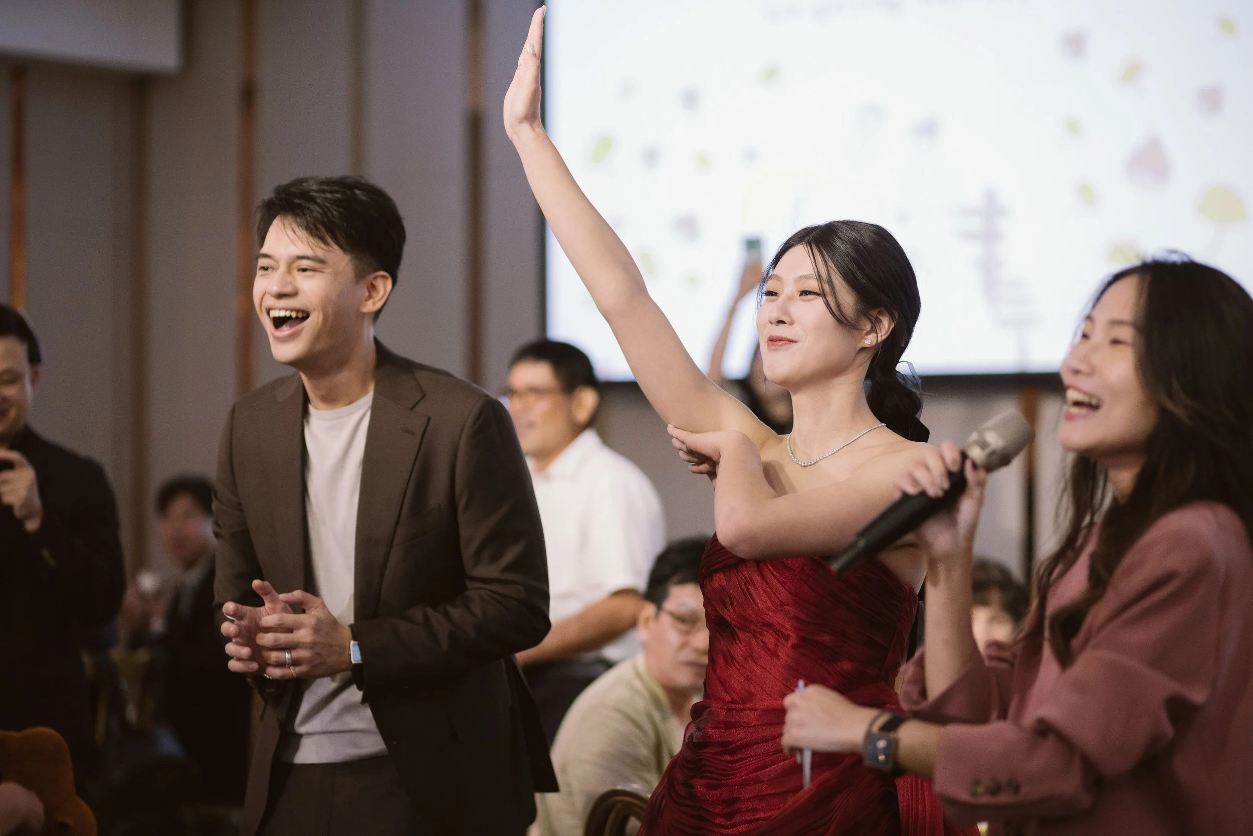 A group of people attending an event, with one woman in a red dress raising her hand. They are smiling, and one person is holding a microphone.