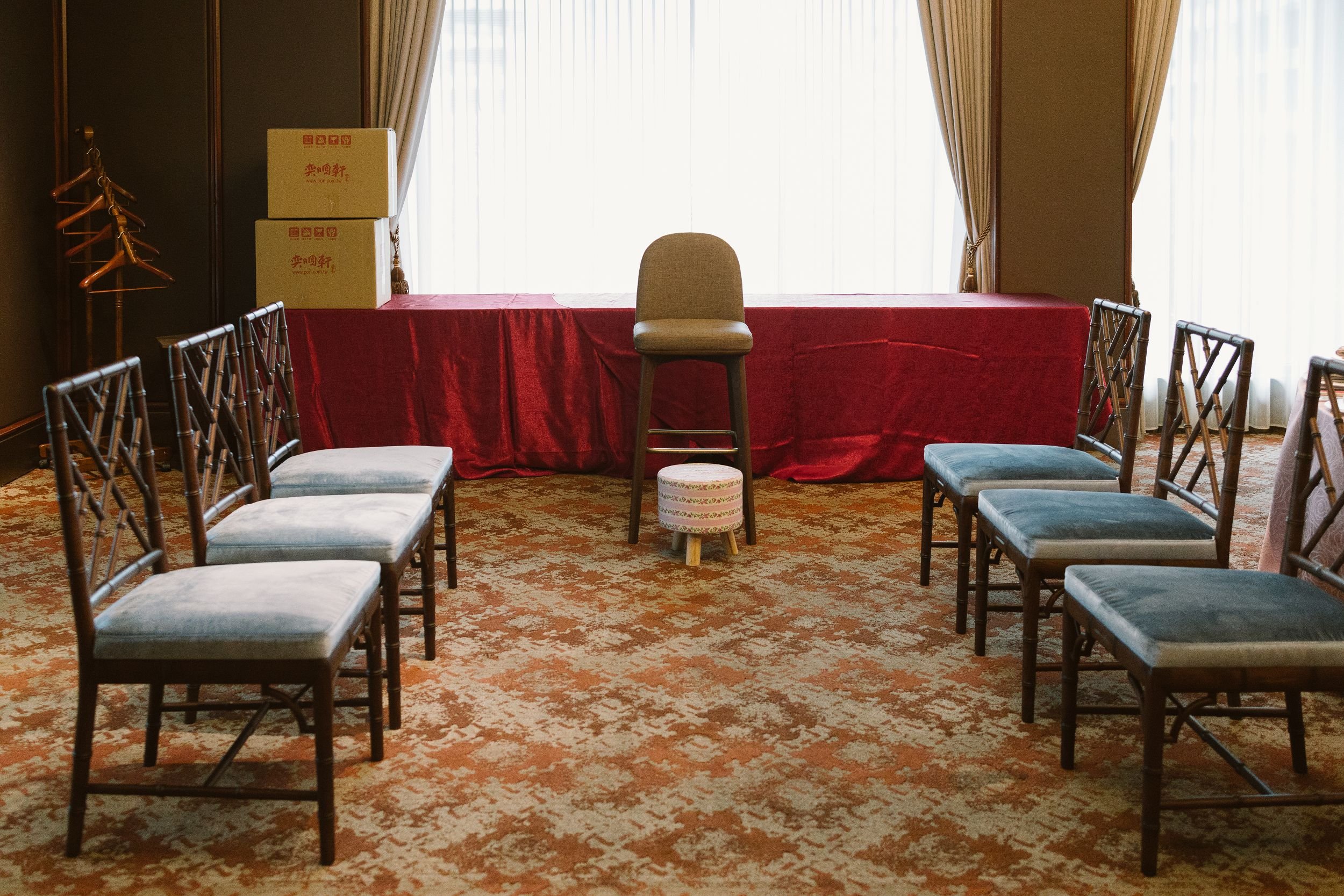 A room with a table covered with a red cloth, surrounded by chairs with cushioned seats. There are potted boxes on the table, a small stool in front of the table, and a coat rack on the left. Large curtains cover windows at the back.