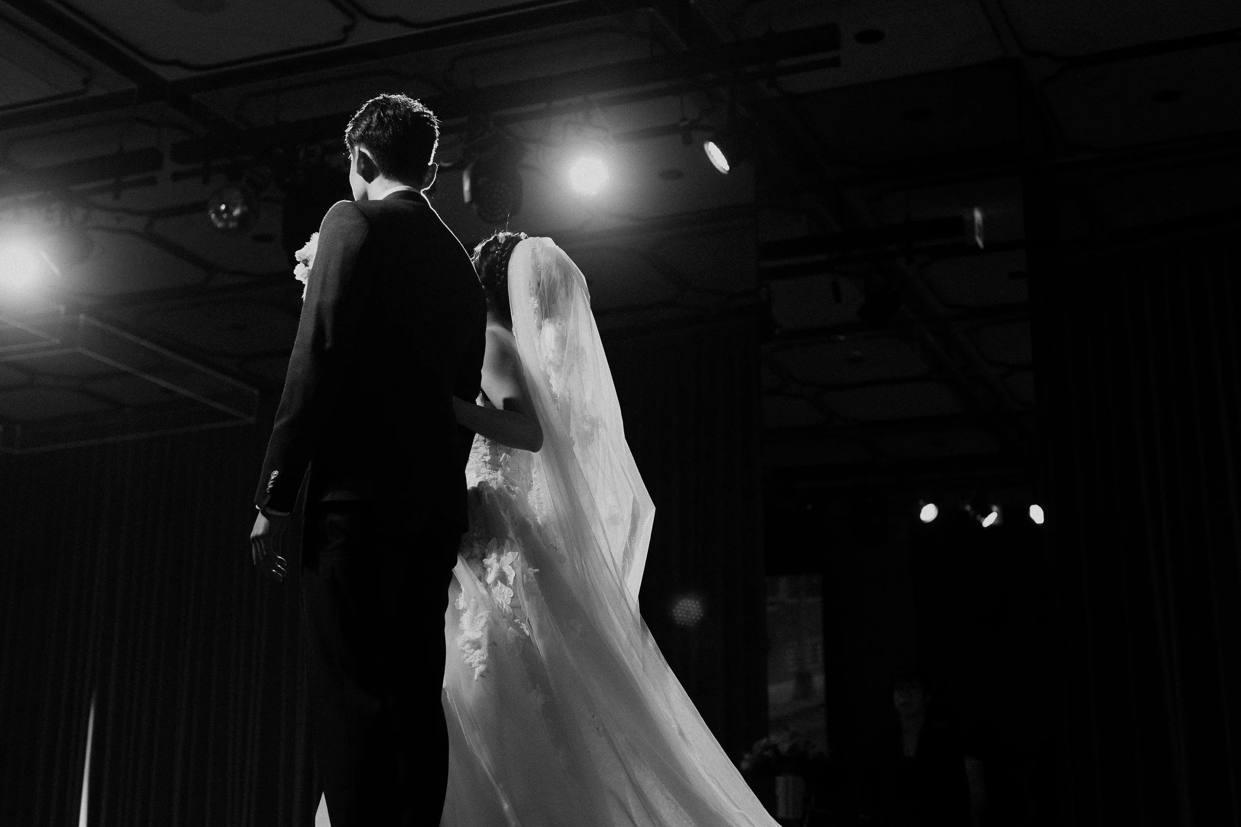 Black and white photo of a bride and groom sharing a dance, with stage lighting overhead and a dark background.