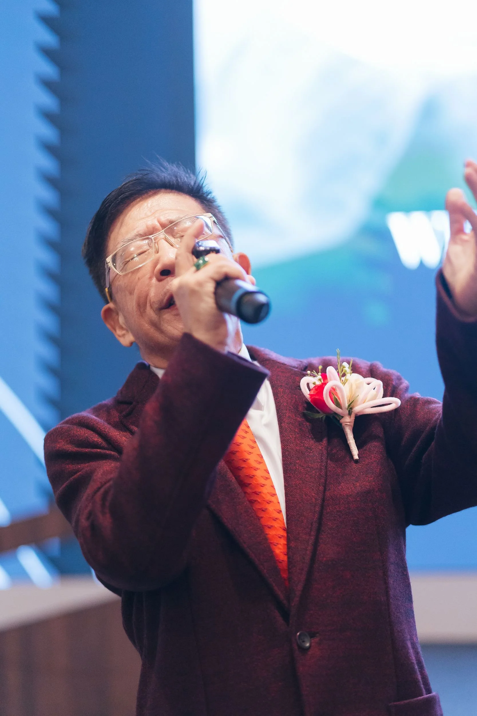 A man singing passionately into a microphone at an indoor event, wearing glasses, a maroon blazer, and a flower boutonniere, with a blue background.