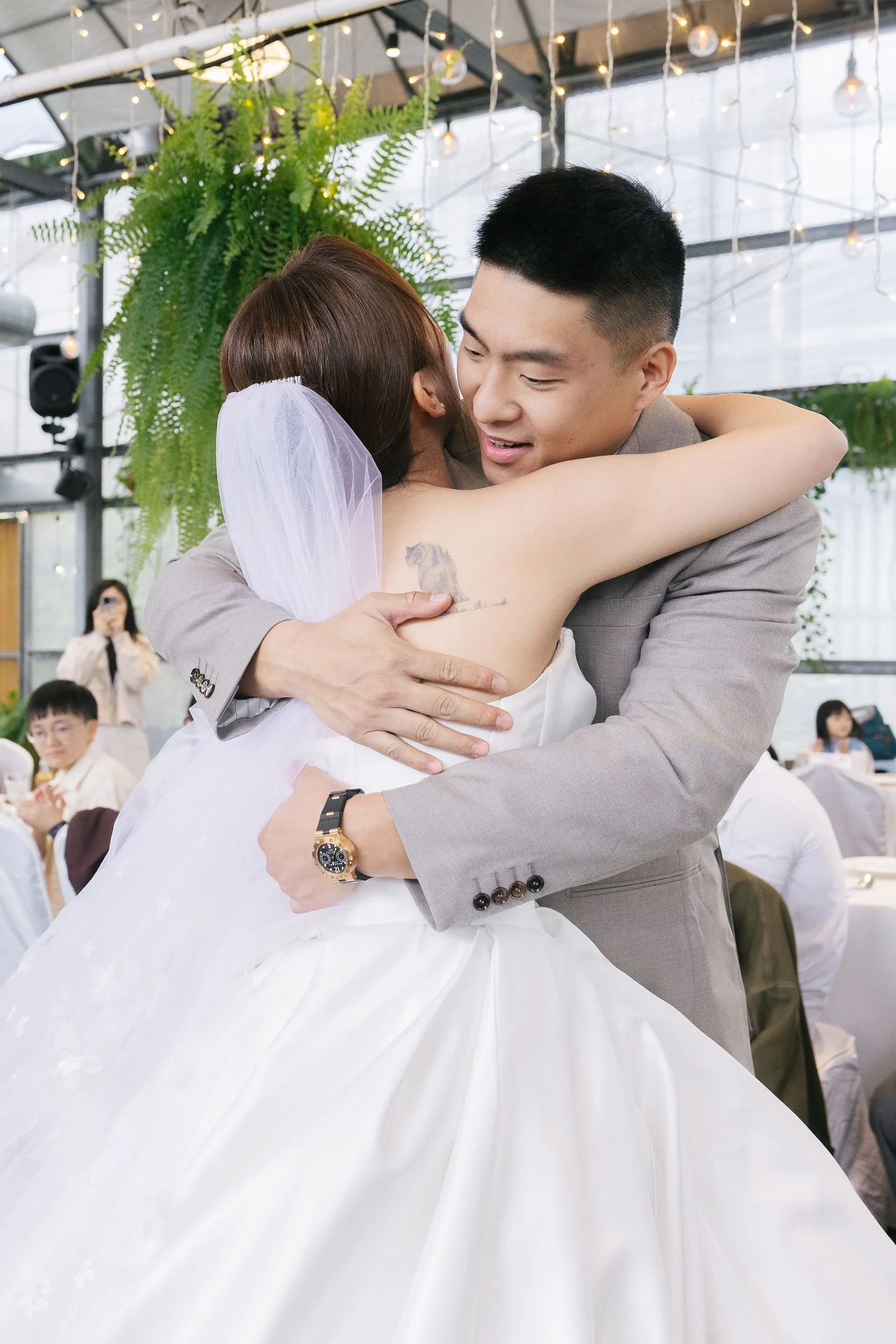 A bride and groom happily hug each other during their wedding reception, with the bride wearing a white wedding dress and veil, and the groom in a gray suit. The background features hanging fairy lights and green plants, with guests seated at tables.
