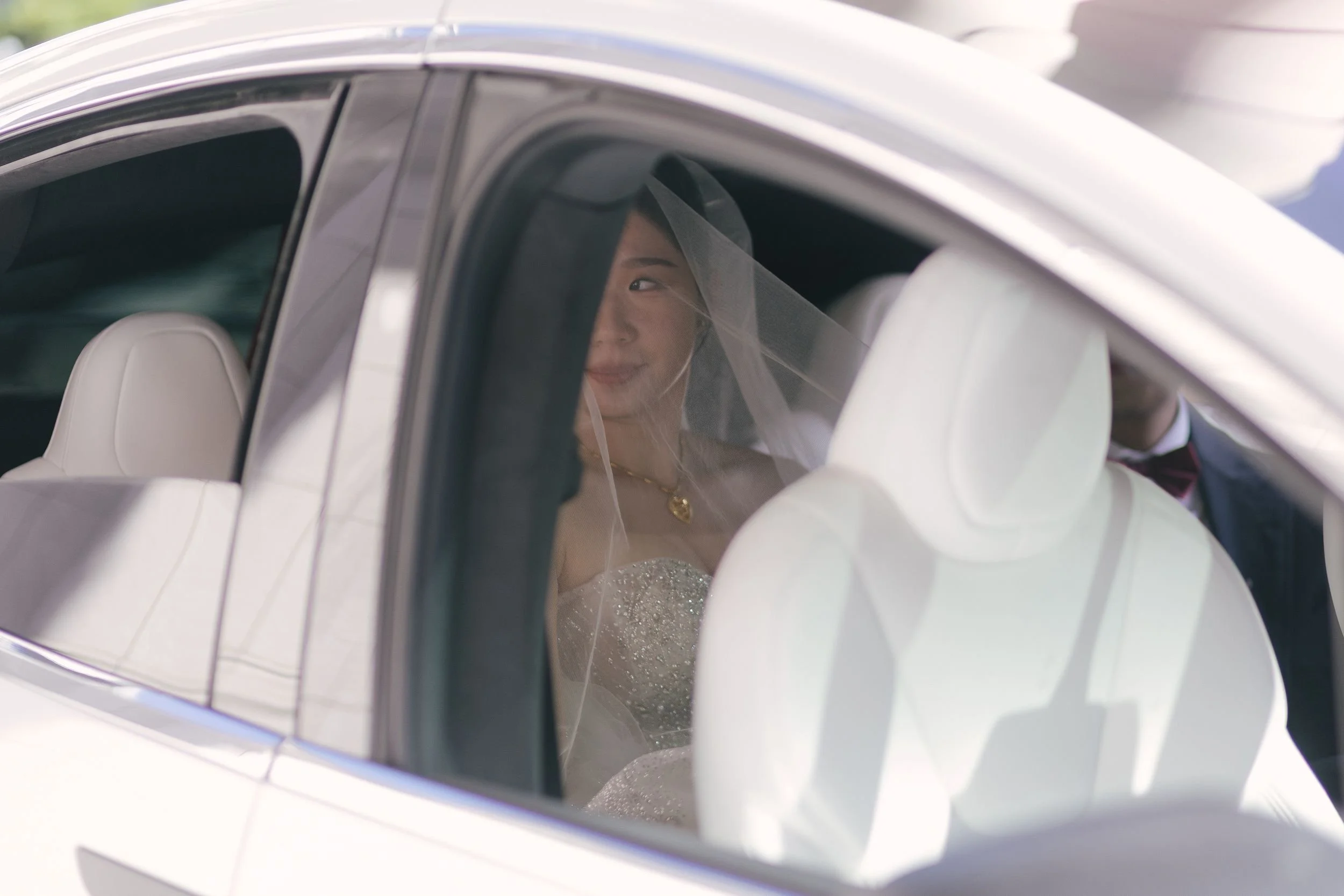A woman dressed in white, wearing a veil and wedding dress, sitting inside a white car, looking out through the window.