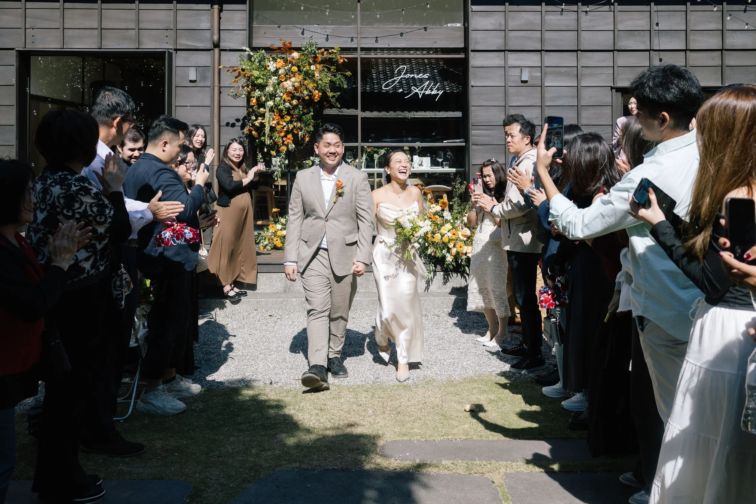A bride and groom walking hand in hand, smiling, as guests gather around them and take photos during an outdoor wedding celebration.