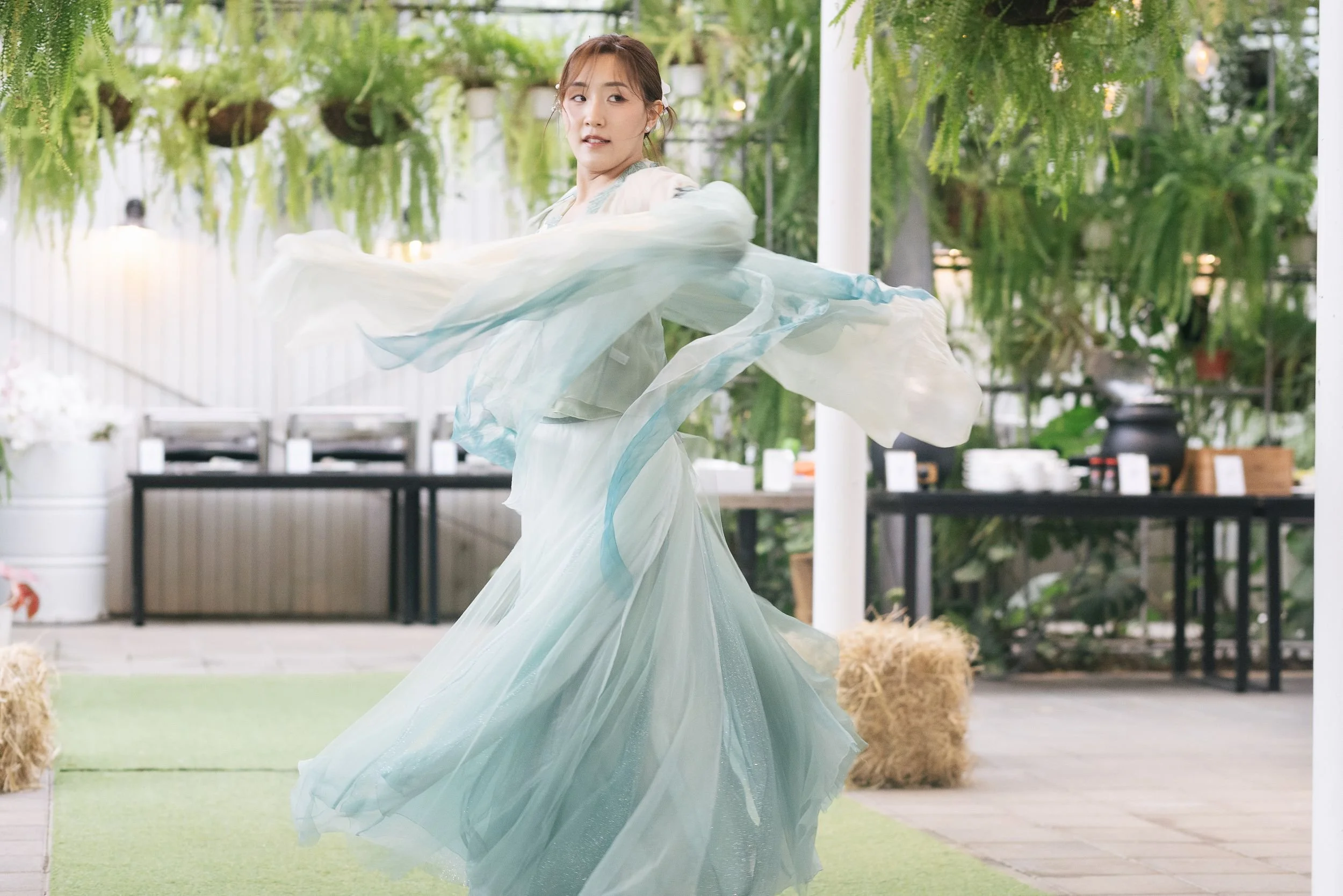 A young woman in a flowing, pastel-colored dress spins gracefully outdoors among lush greenery and hanging plants.