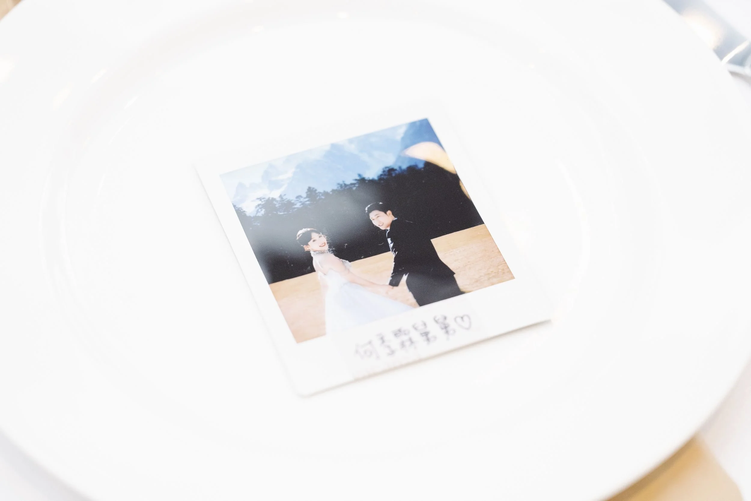 A printed photo of a smiling couple holding hands outdoors at sunset, with mountains and trees in the background.