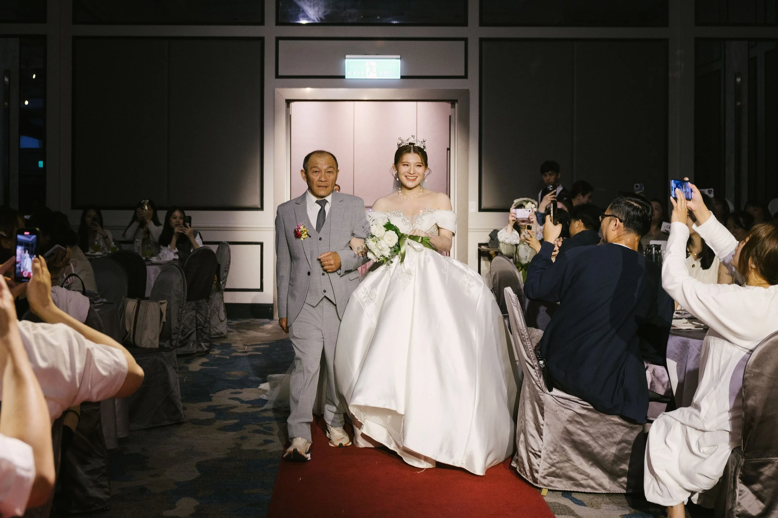 A bride in a wedding gown smiling and walking arm-in-arm with an older man in a suit, in a wedding reception setting with guests taking photos.