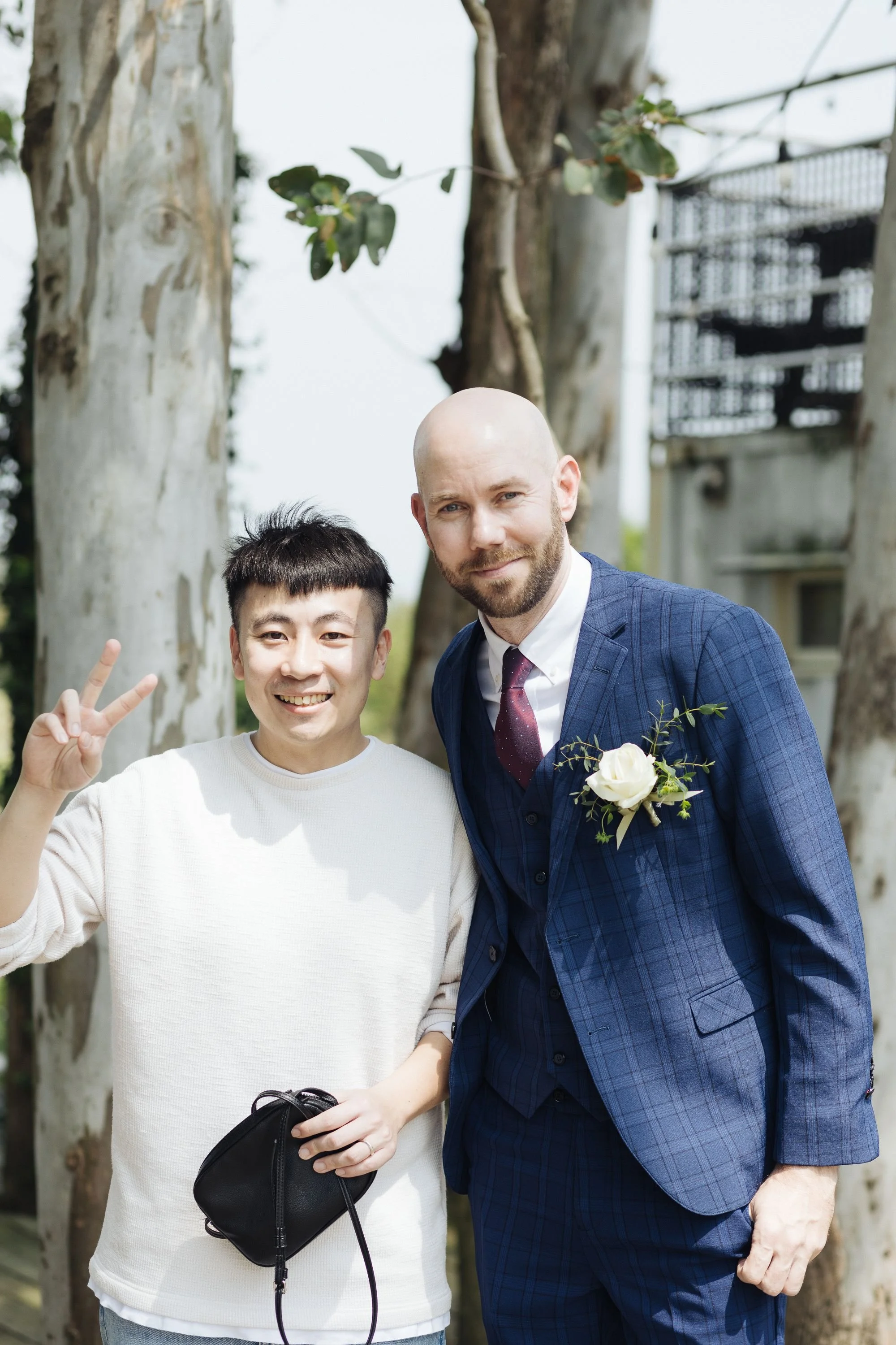 Two men smiling outdoors in front of trees; one is young, holding a small black bag, and the other is older, dressed in a blue plaid suit with a white flower boutonniere.