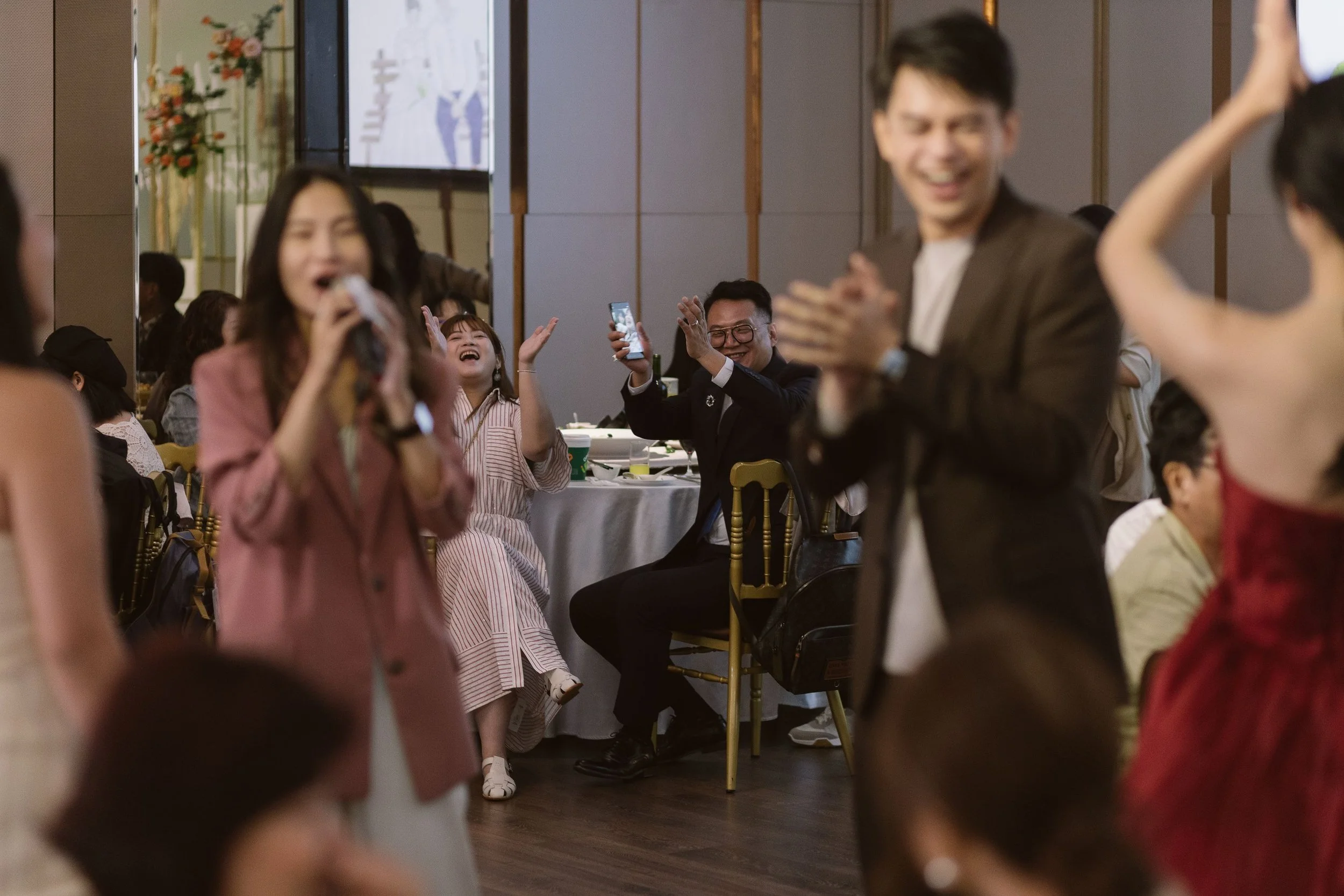 People laughing and enjoying a celebration, with some taking photos, in a decorated indoor venue.